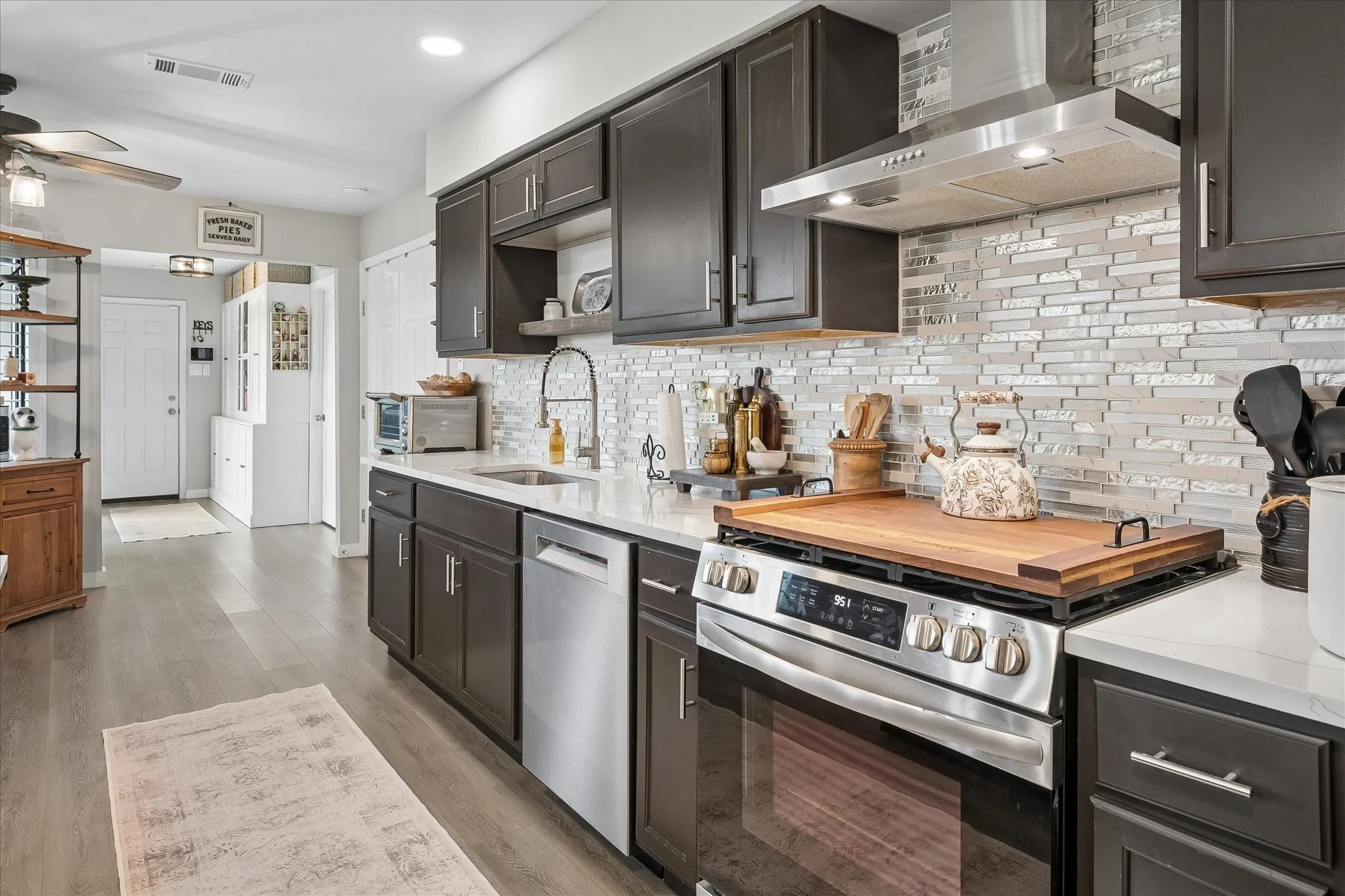 Kitchen featuring dark cabinetry, solid-surface countertops, horizontal glass-tile backsplash, stainless steel appliances, and wood-finish flooring