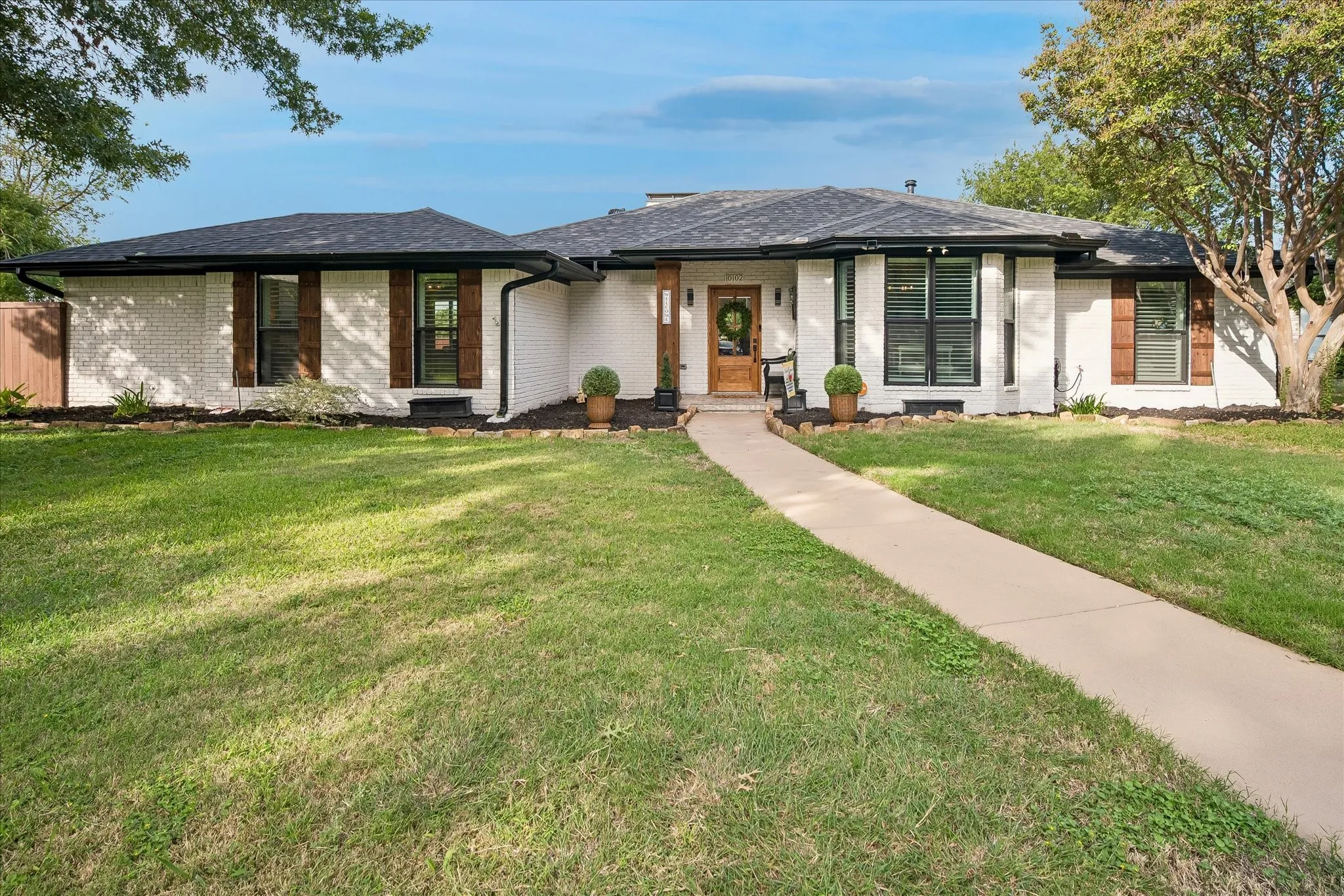 Single-story exterior featuring painted brick masonry, dark-finish roof shingles, decorative wood-finish shutters, a covered entry with a timber-style column, and a concrete walkway