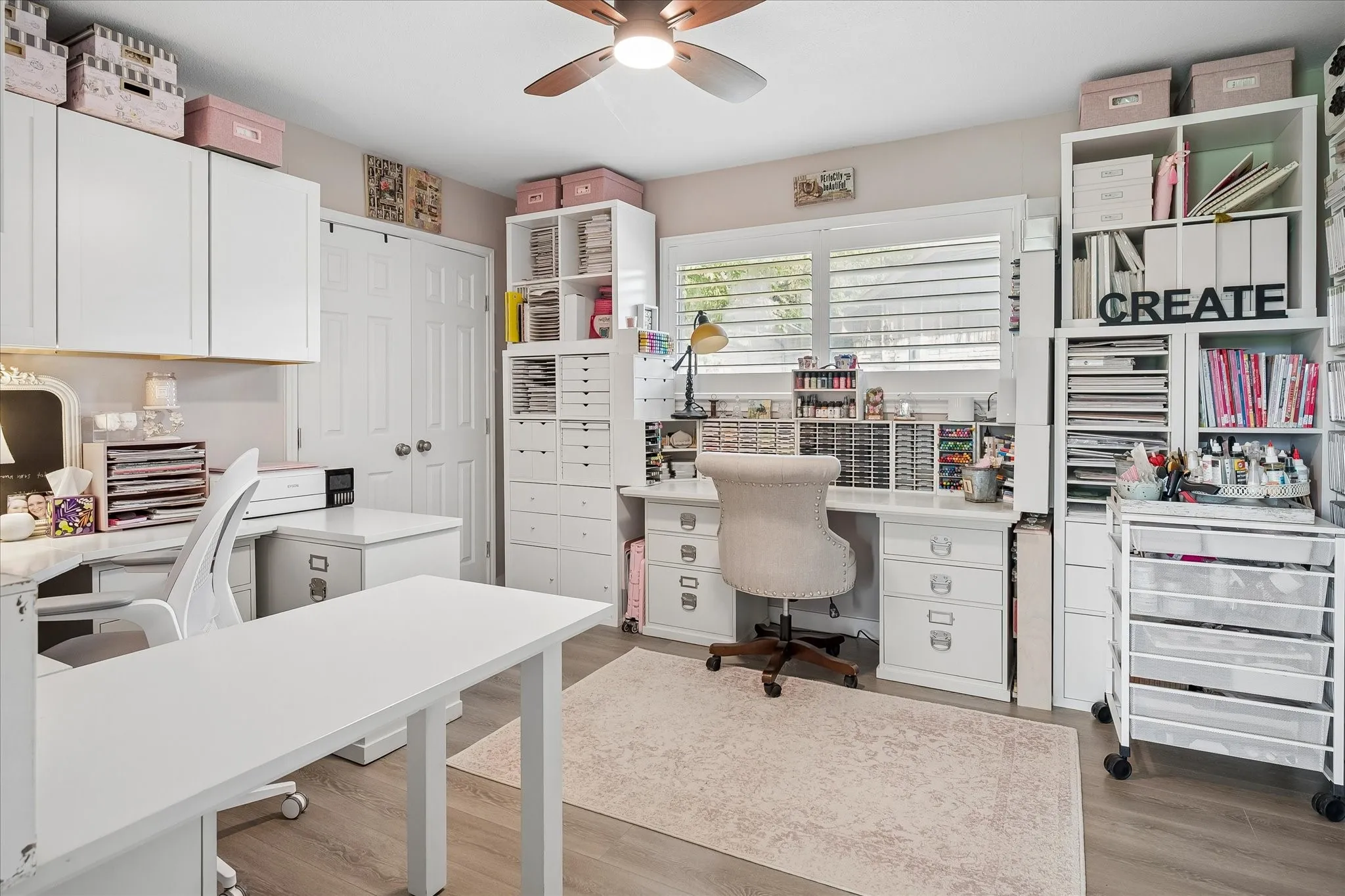 Custom built-in white cabinetry, integrated desk workspace, wood-finish flooring, ceiling fan, and window shutters