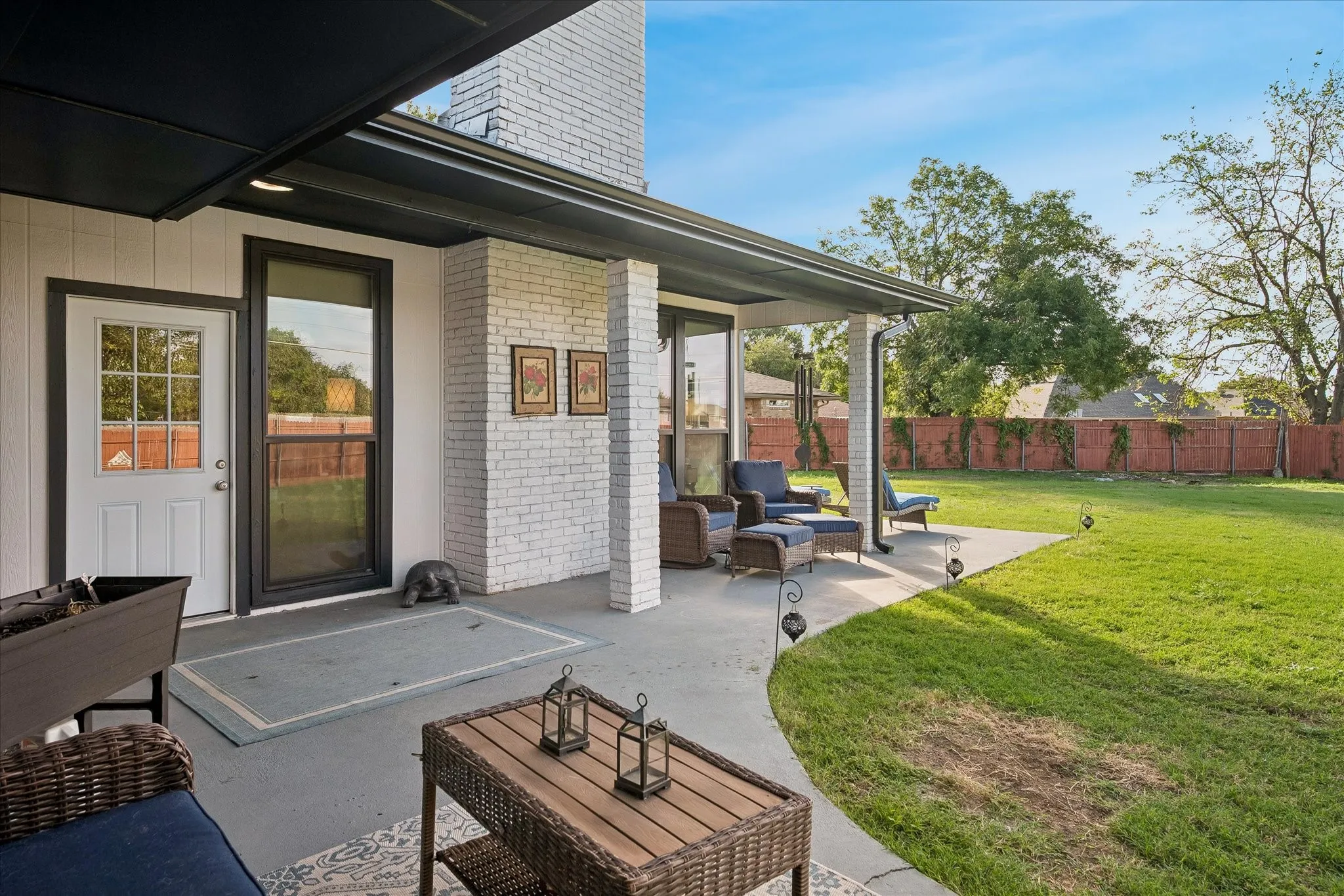 Covered concrete patio featuring a white brick masonry pillar, exterior wall-mounted lighting, and a spacious backyard enclosed by wood fencing