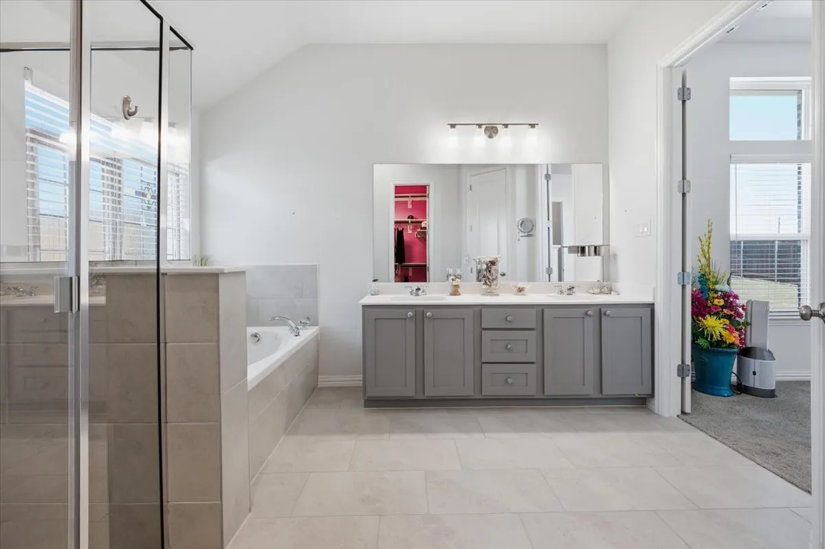 Spacious bath featuring a dual vanity with gray cabinetry, an oversized mirror, a built-in soaking tub, and a separate glass-enclosed shower