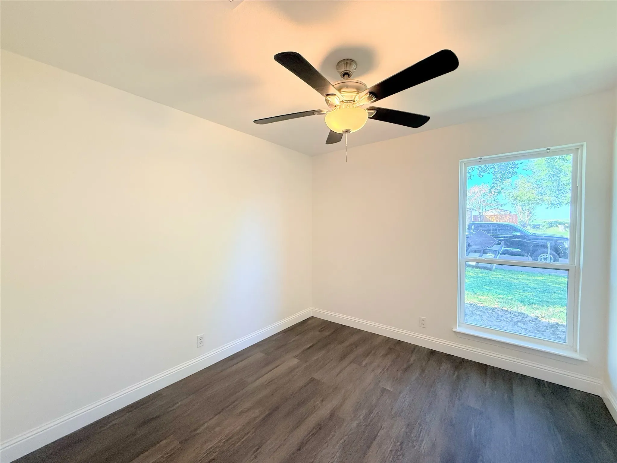 Bright bedroom featuring dark wood-finish flooring, a ceiling fan with integrated lighting, and a large window with natural light exposure