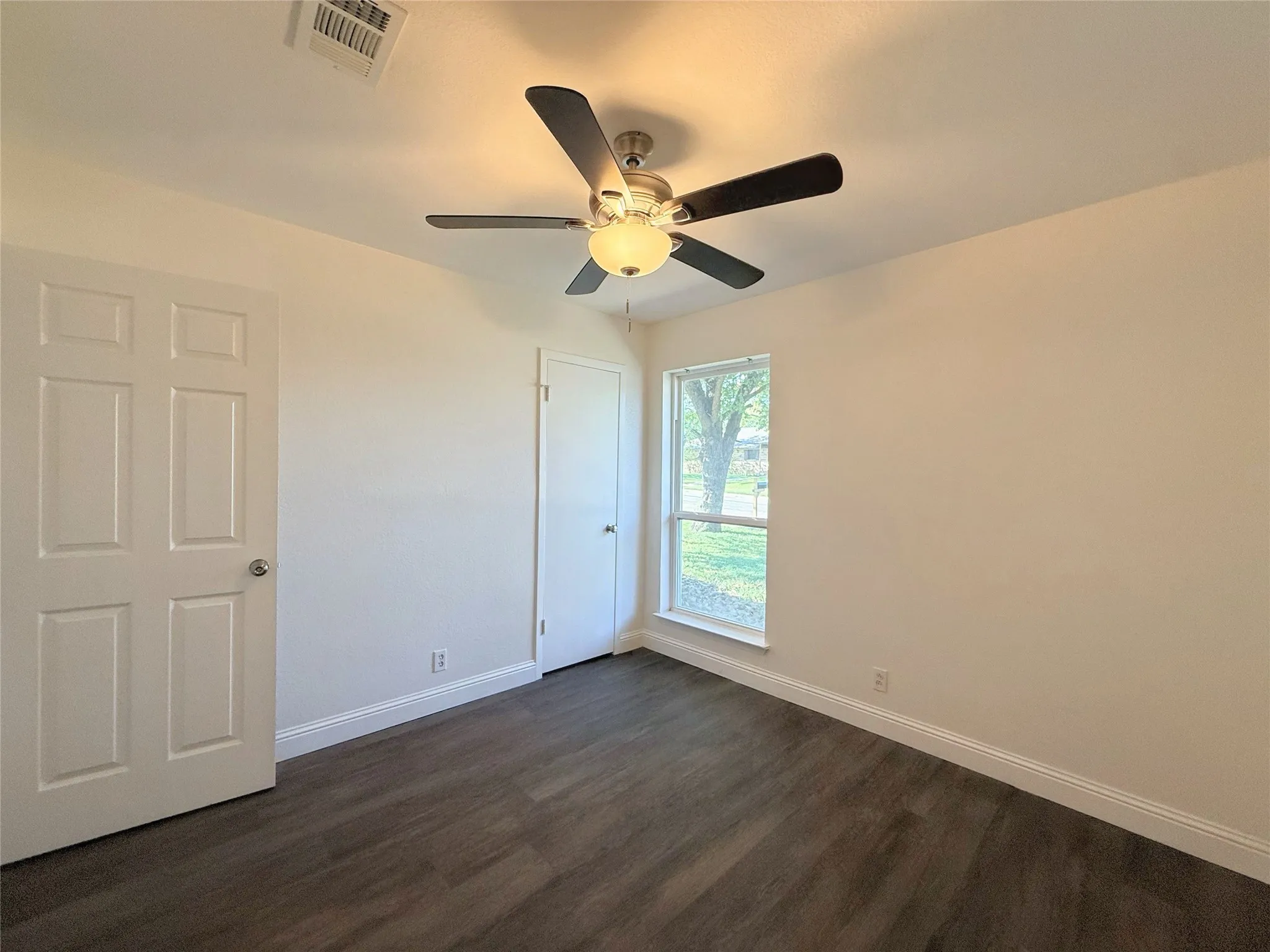 Bright bedroom featuring dark wood-finish flooring, ceiling fan with integrated lighting, and natural light from large window