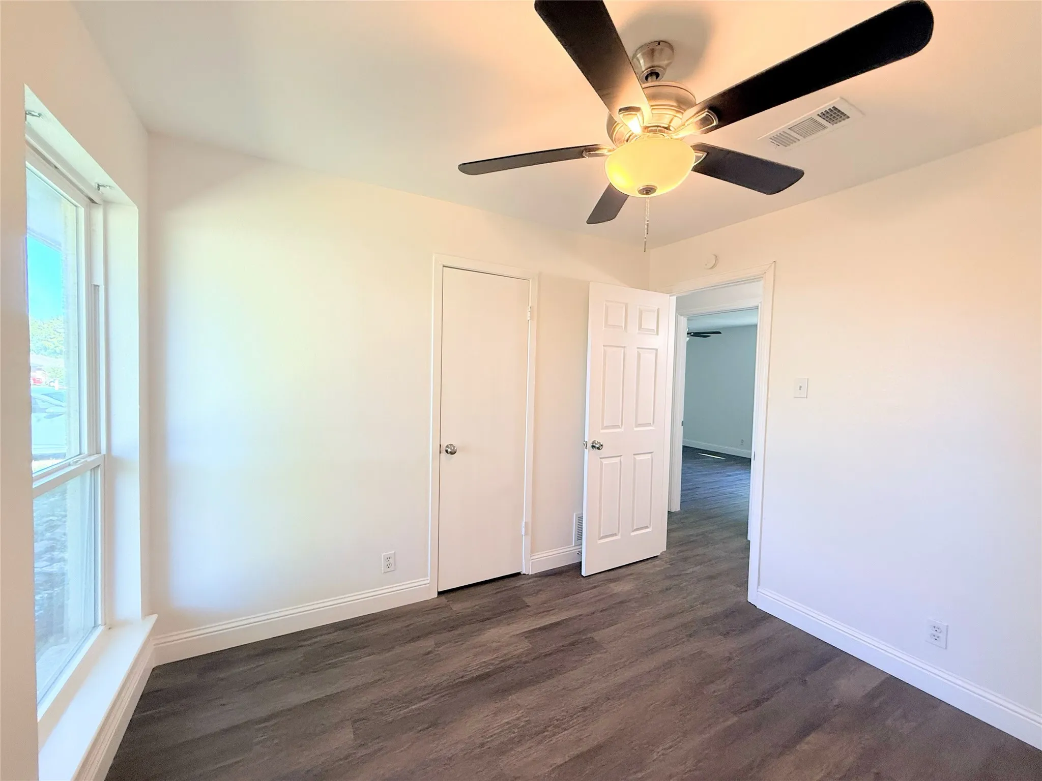 Spacious bedroom featuring dark wood-finish flooring, large windows with natural light, ceiling fan with integrated lighting, and multiple closet doors for storage