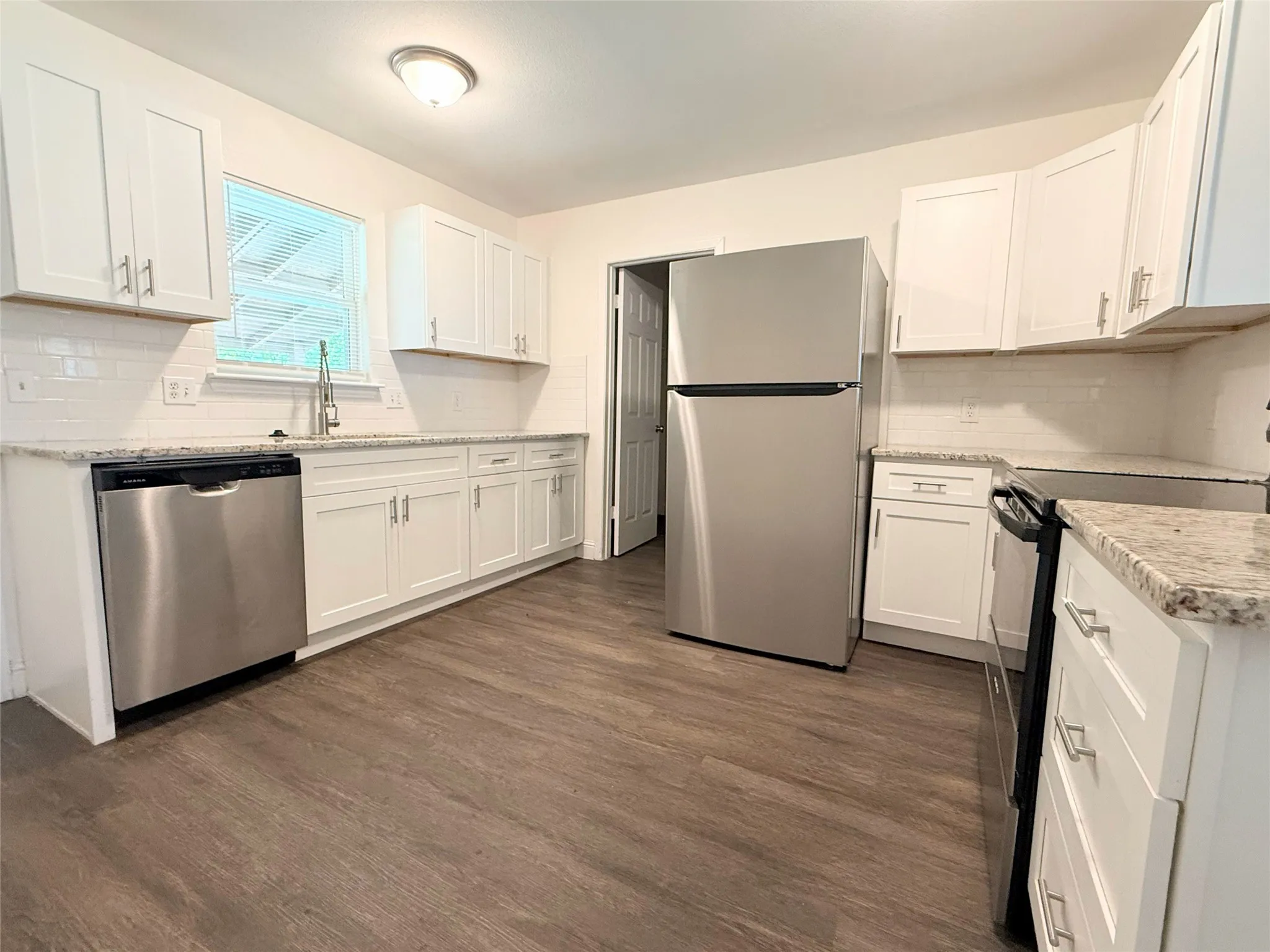 Bright, updated kitchen featuring white cabinetry with brushed nickel hardware, stainless steel appliances including a refrigerator and dishwasher, wood-finish flooring, subway tile backsplash, light countertops, and a natural light window overlooking the grounds