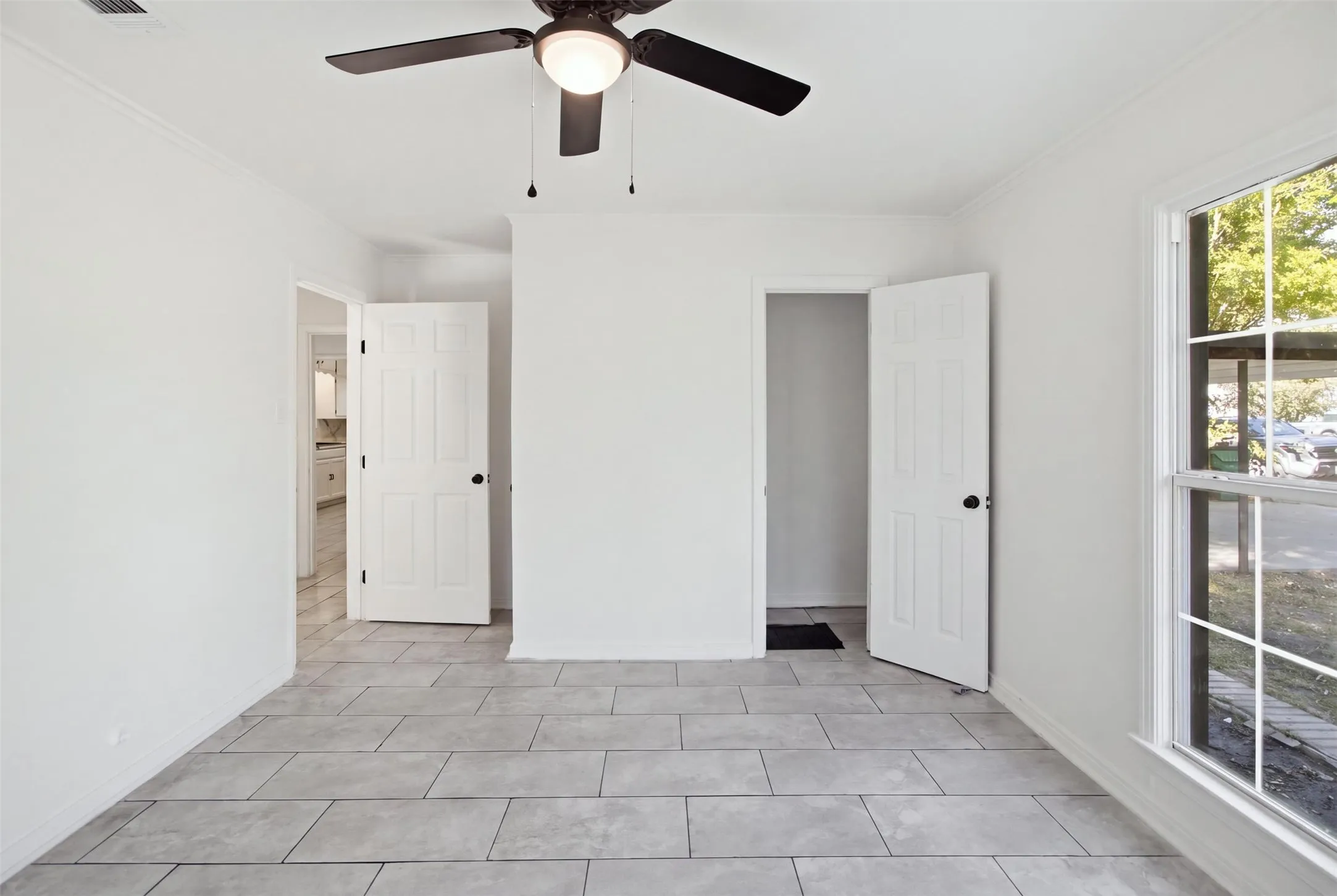 Light-toned rectangular tile flooring, white interior doors with black hardware, a ceiling fan with integrated lighting, and a multi-pane window