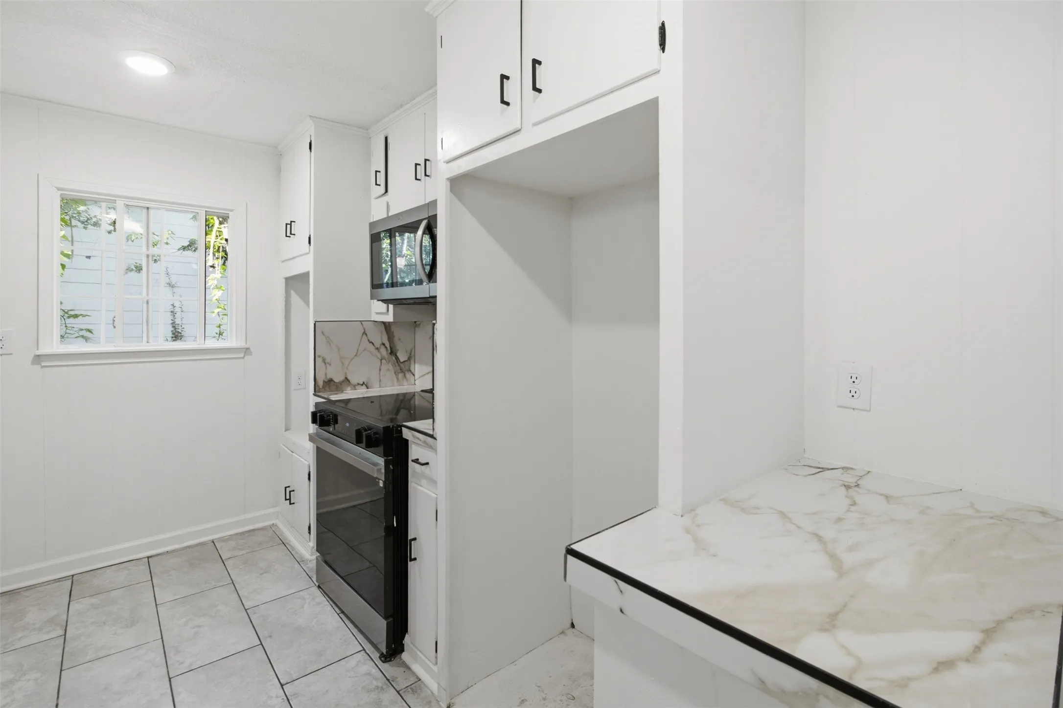 White kitchen cabinetry with black hardware, stainless steel range and microwave, marbled countertops, and tile flooring