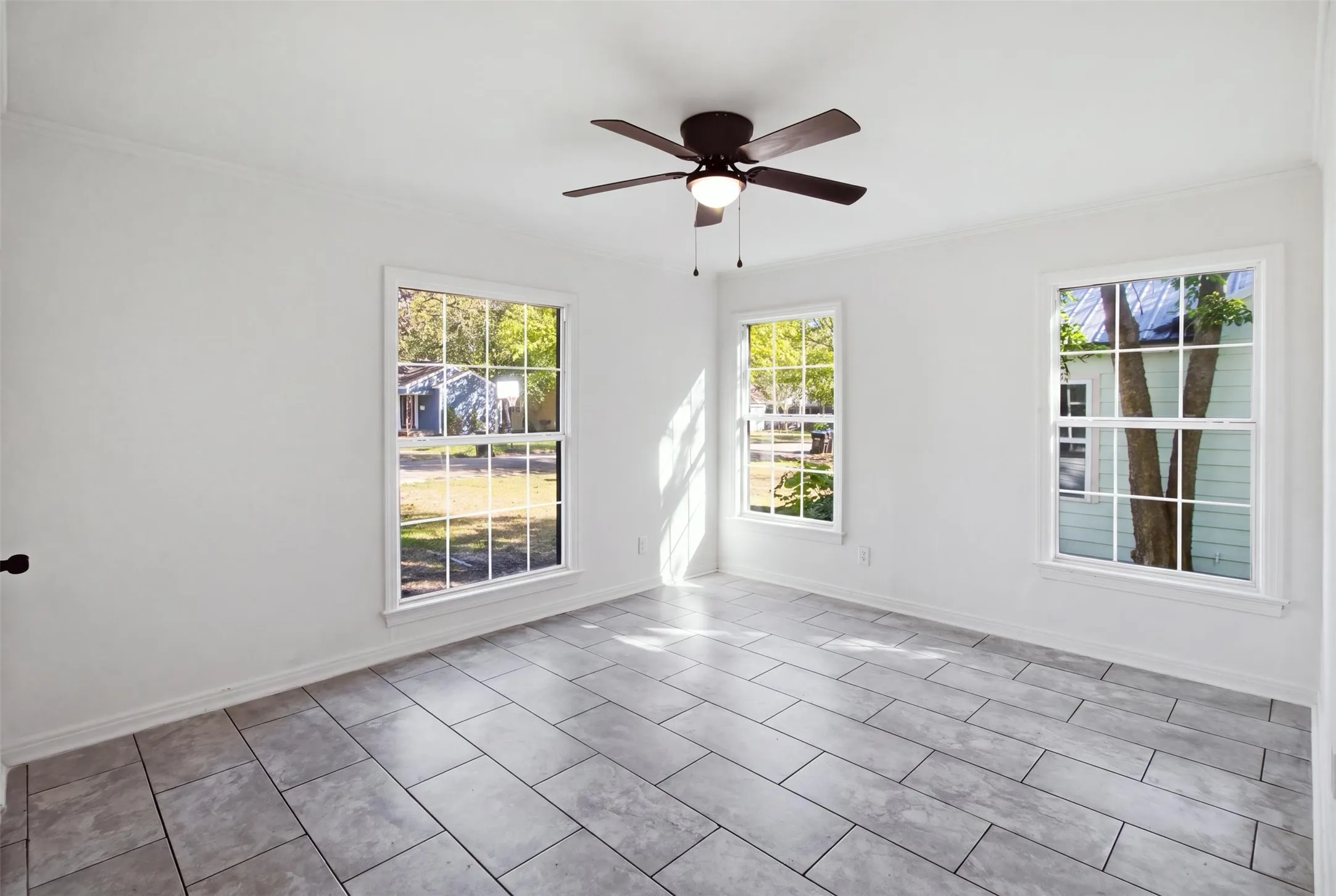 Light-filled room featuring three large windows, a ceiling fan with integrated lighting, crown molding, and large format tile flooring