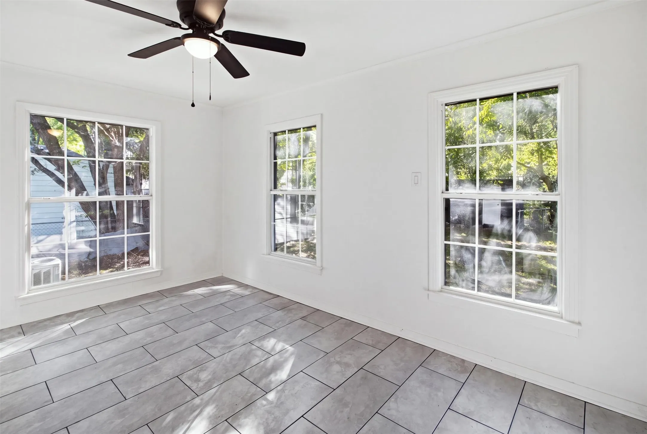 Three bright windows with grid panes, a ceiling fan with light, and light-toned rectangular floor tiles
