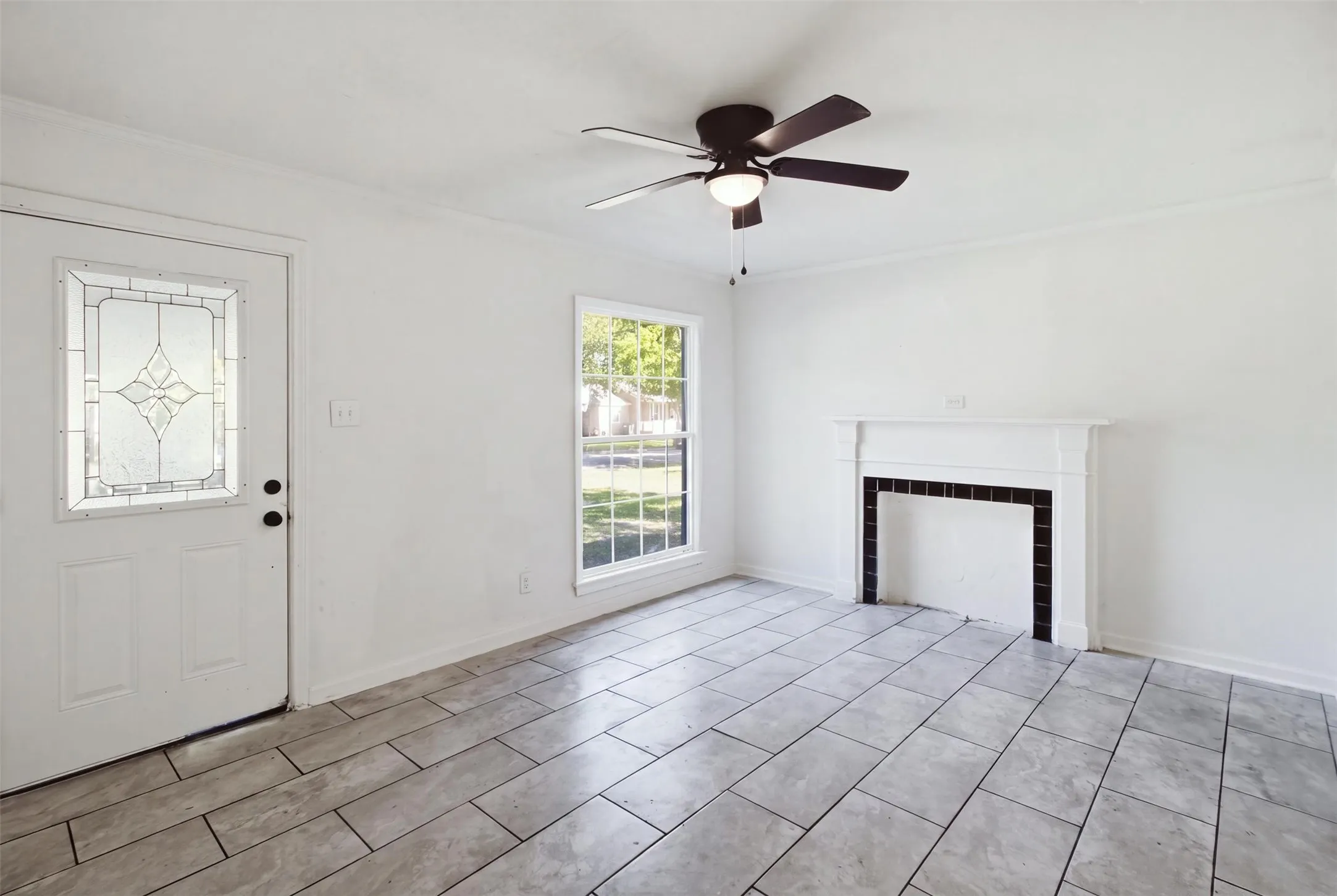 Entry door with decorative glass insert, ceiling fan with integrated lighting, a single window, white walls, and rectangular tile flooring