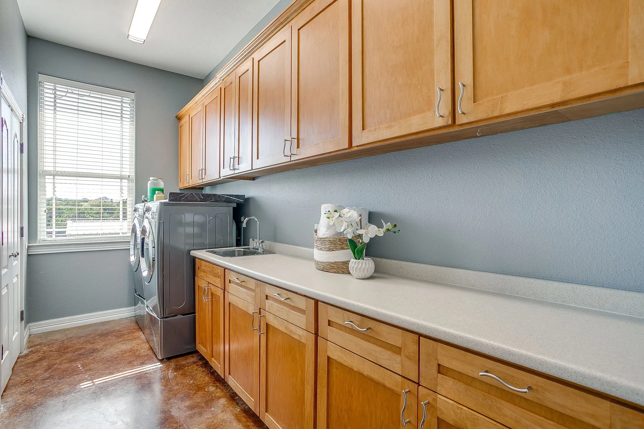 Light-filled laundry room featuring extensive wood cabinetry, a large window, a utility sink, and ample countertop space