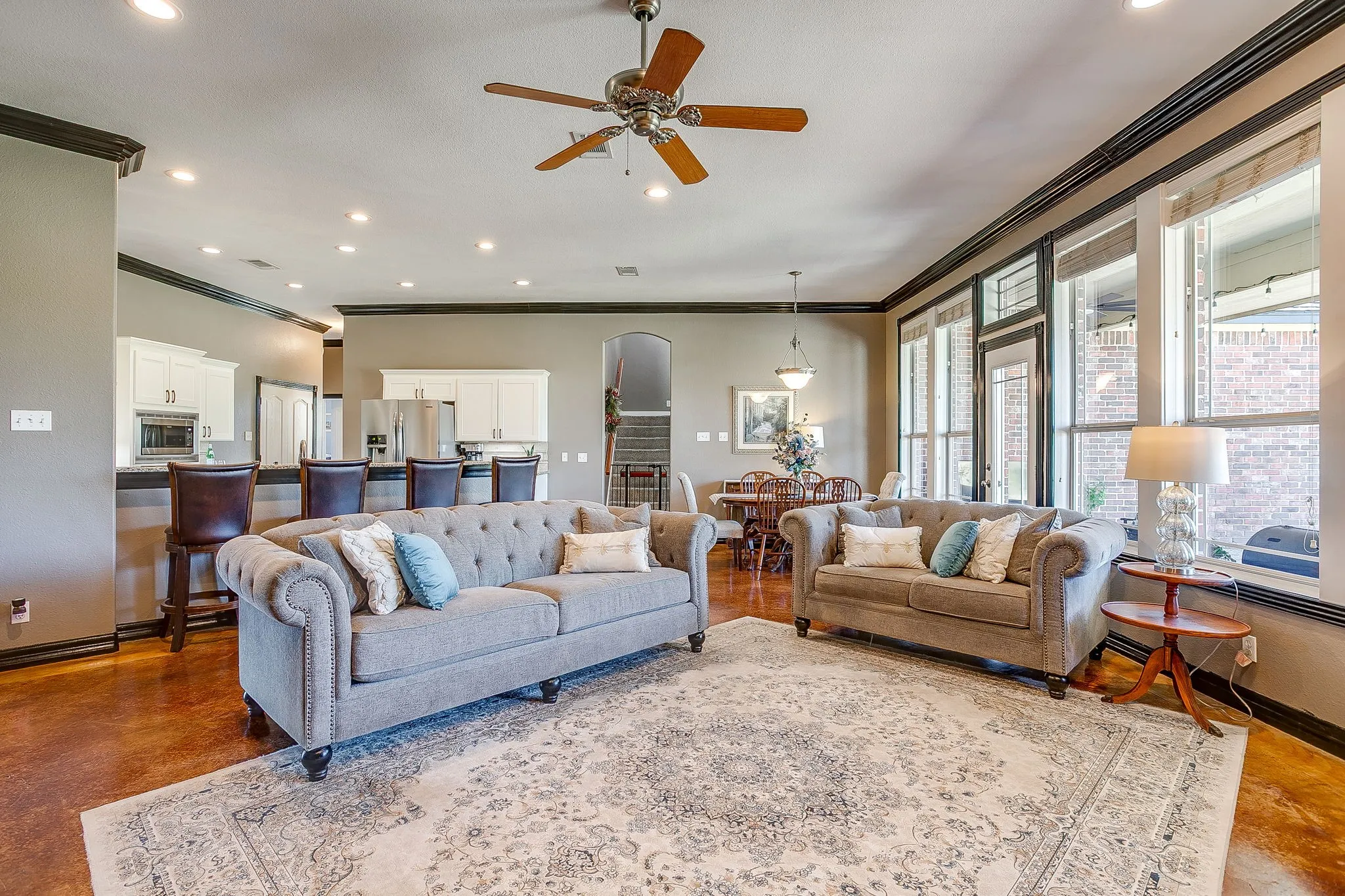 Open-concept living area featuring hardwood flooring, a coffered ceiling with recessed lighting and ceiling fan, large multi-pane windows with natural light, and an adjacent kitchen with stainless steel appliances and white cabinetry