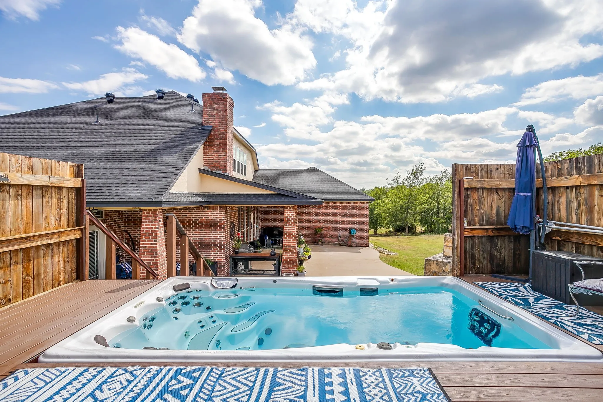 Expansive composite deck with composite decking and integrated outdoor living spaces, featuring an in-ground swimming pool with spa jets, mature landscape backdrop with established trees, brick chimney detail on the home exterior, and ample seating and dining areas with patio furnishings