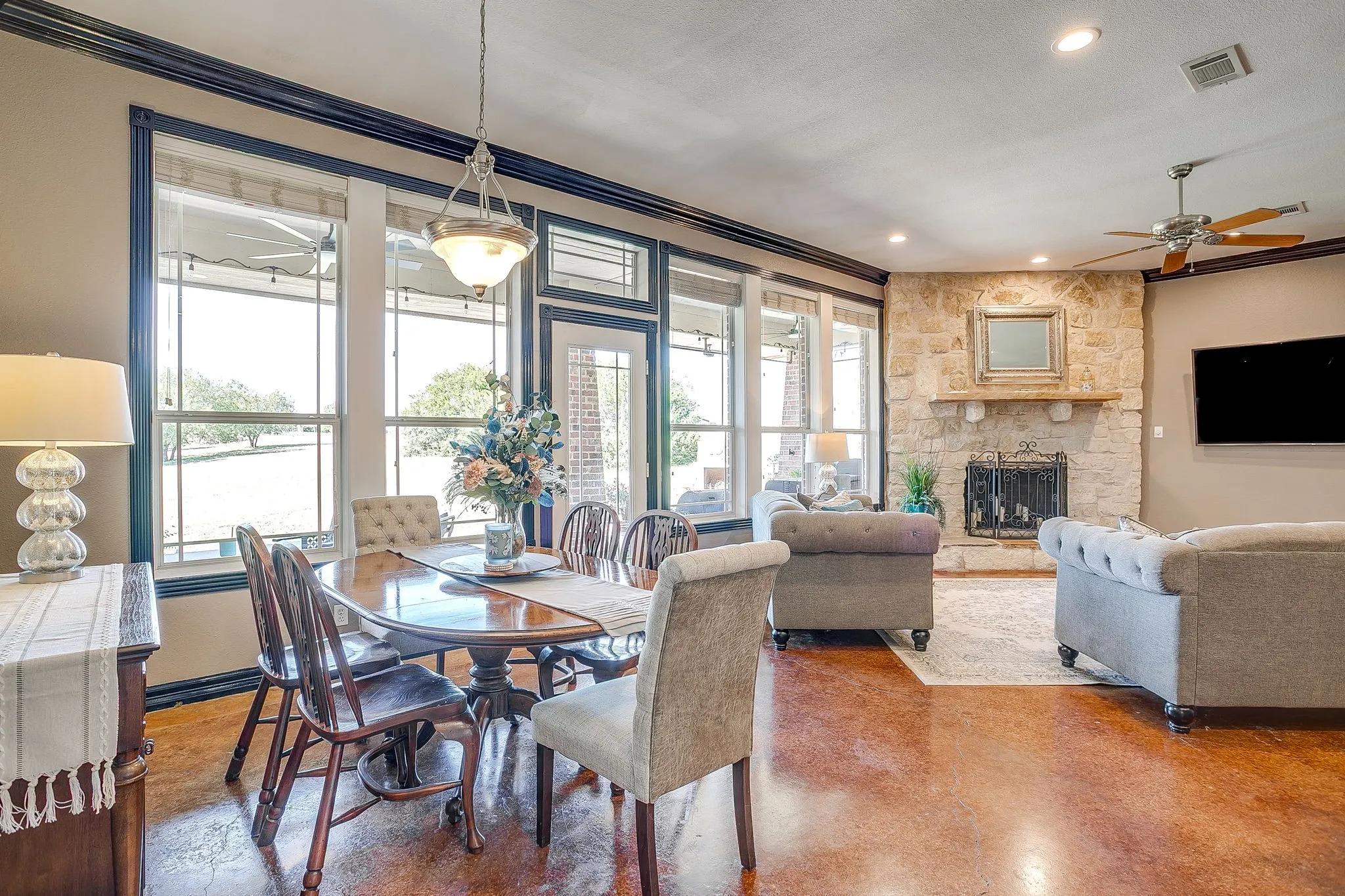 Open-concept living area featuring expansive multi-panel glass doors and windows with dark frames, wood-finish flooring throughout, a dining table with upholstered chairs, built-in fireplace with stone surround, ceiling fan, recessed lighting, and a wall-mounted flat-screen television