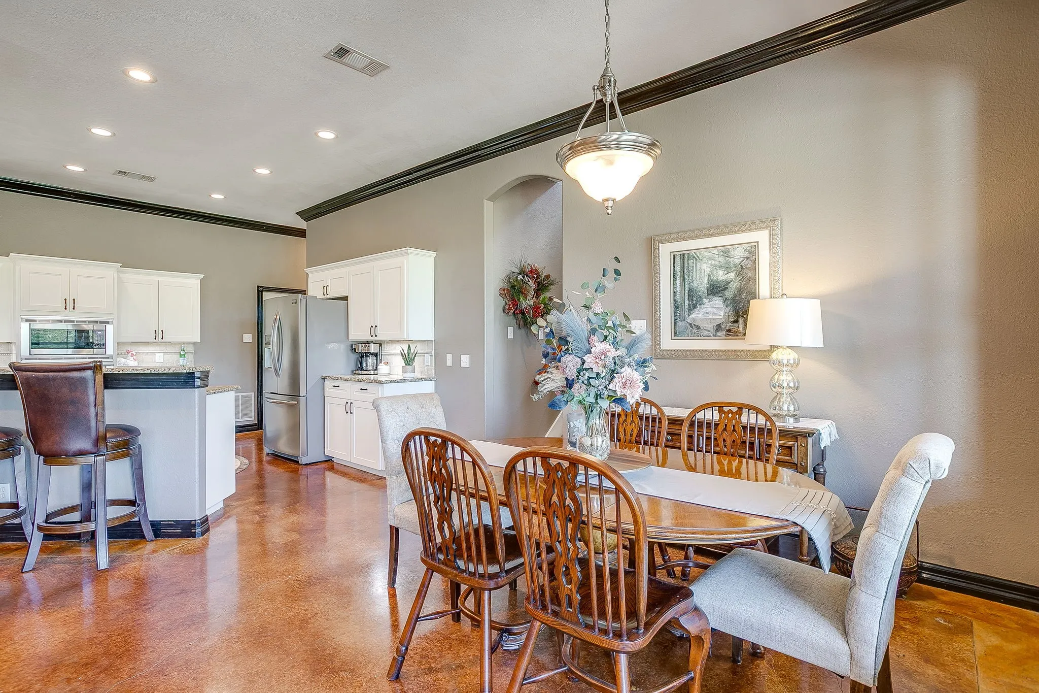 Open-concept dining and kitchen area featuring wood-finish flooring, white cabinetry with stainless steel appliances, exposed ceiling beams, recessed lighting, and a pendant fixture above the dining table with seating for six