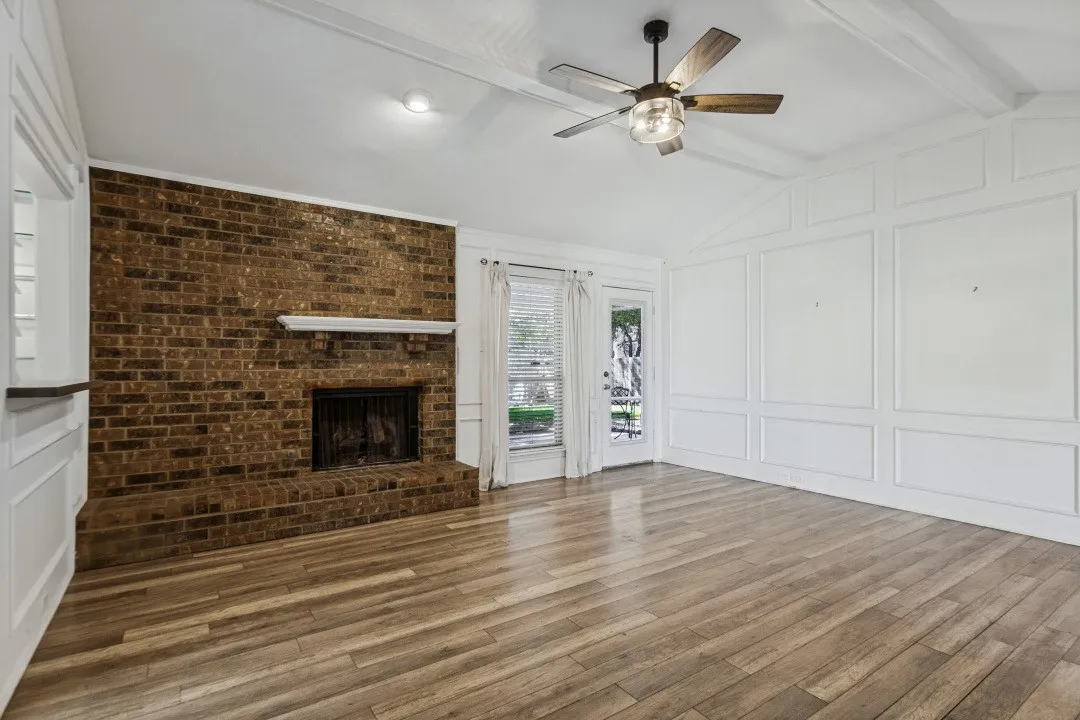 Spacious open-concept living area featuring a striking exposed brick fireplace with mantel, warm wood-finish flooring throughout, recessed lighting, ceiling fan with integrated light fixture, and glass-paneled French doors providing natural light and access to outdoor space