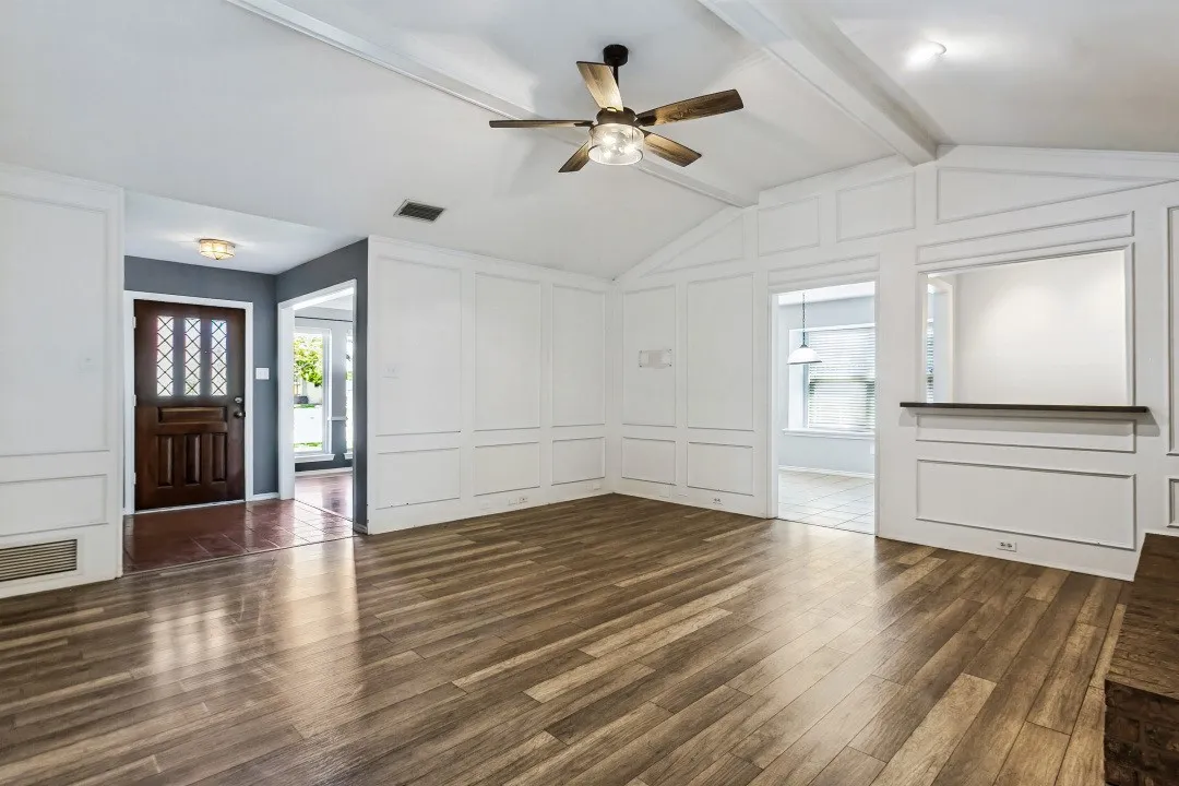 Vaulted ceiling with exposed beam, wood-finish flooring, decorative wall paneling, built-in display shelf, and an entry door with decorative glass inserts