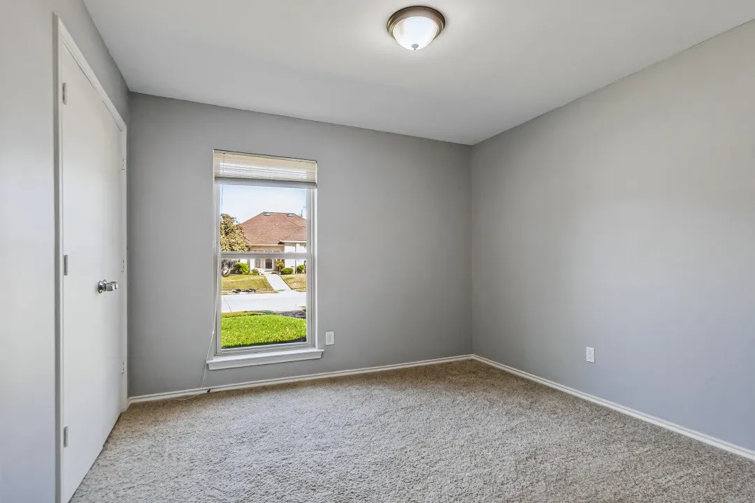 Light gray wall color, carpeted flooring, single window with blinds, flush-mount ceiling light, white trim throughout
