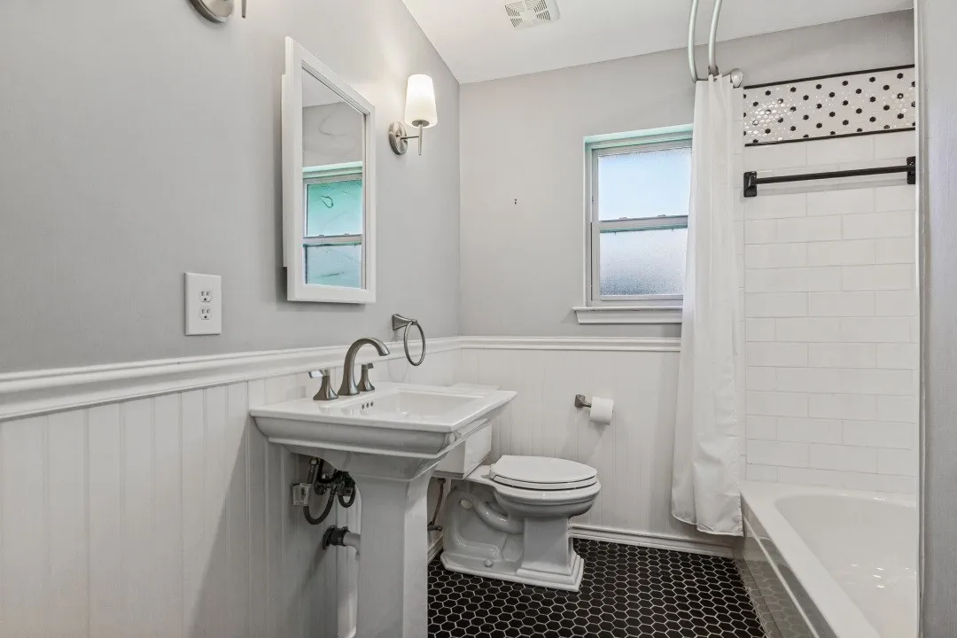 Pedestal sink with chrome fixtures, white wainscoting, black hexagonal floor tiles, subway tile shower surround, and a decorative tile accent