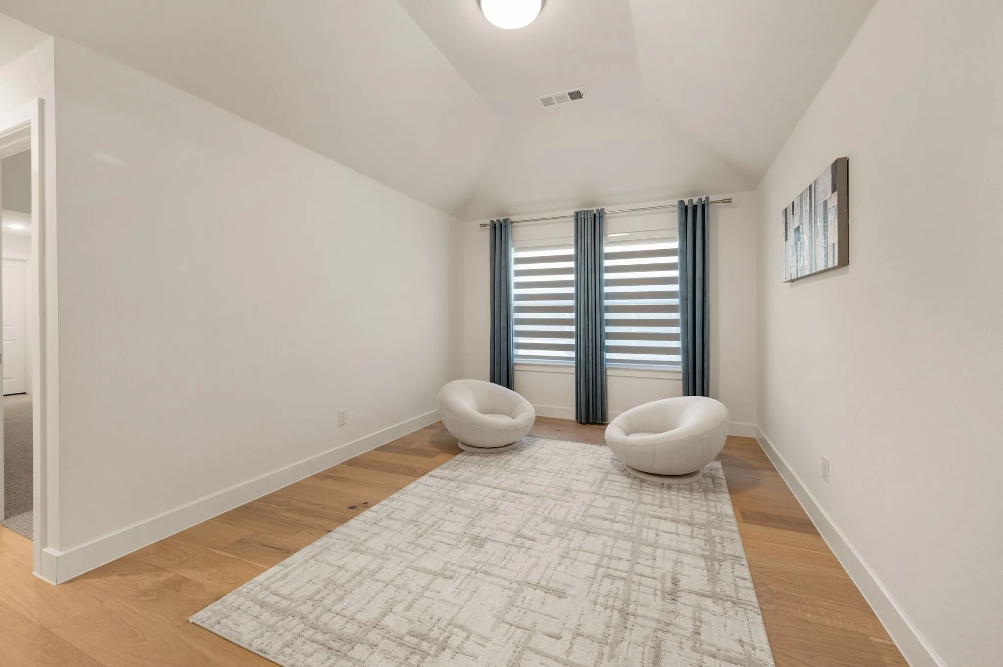 Light wood-finish flooring with white baseboards, two windows with blinds and curtains, and a vaulted ceiling with a flush-mount light fixture