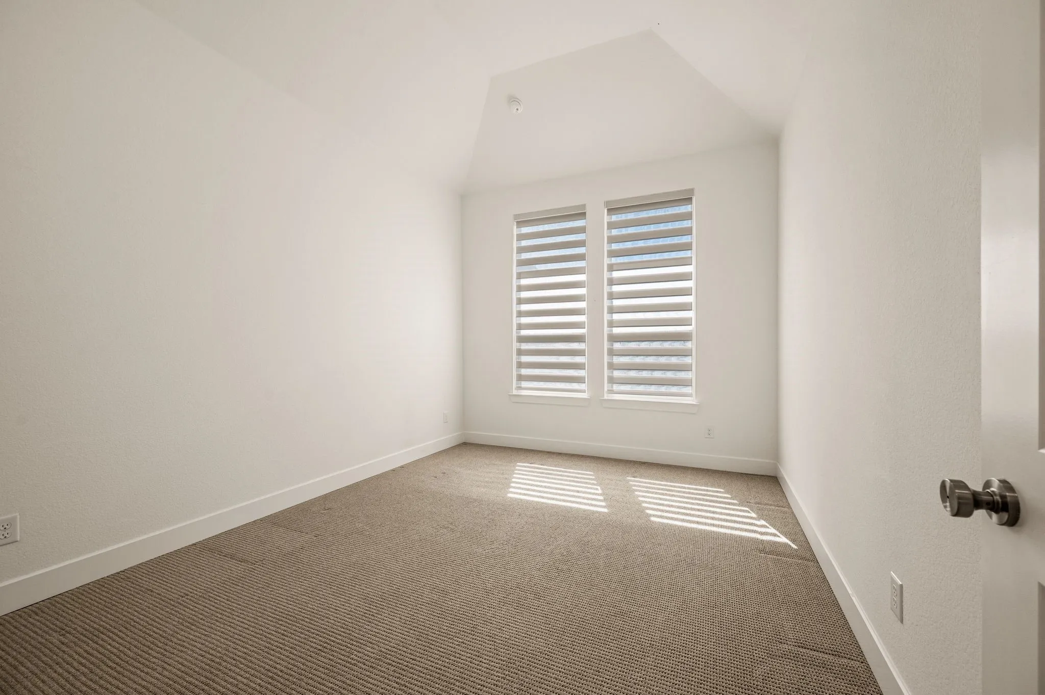 Downstairs Guest Bedroom with vaulted ceiling, two windows with horizontal blinds, neutral-toned carpet, and a white interior door with a dark knob