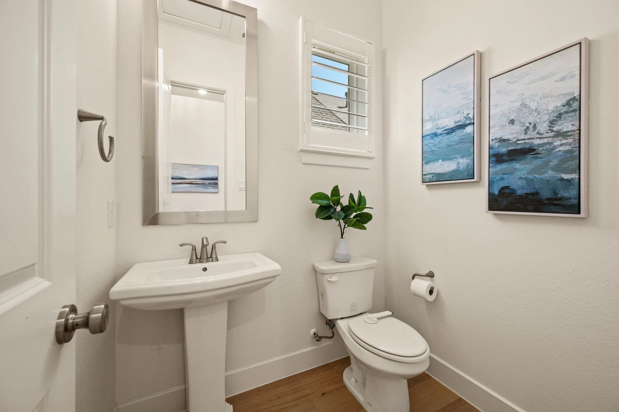 Powder bath with pedestal sink, brushed nickel faucet, mirror with a brushed metal frame, white window shutters, and wood-finish flooring