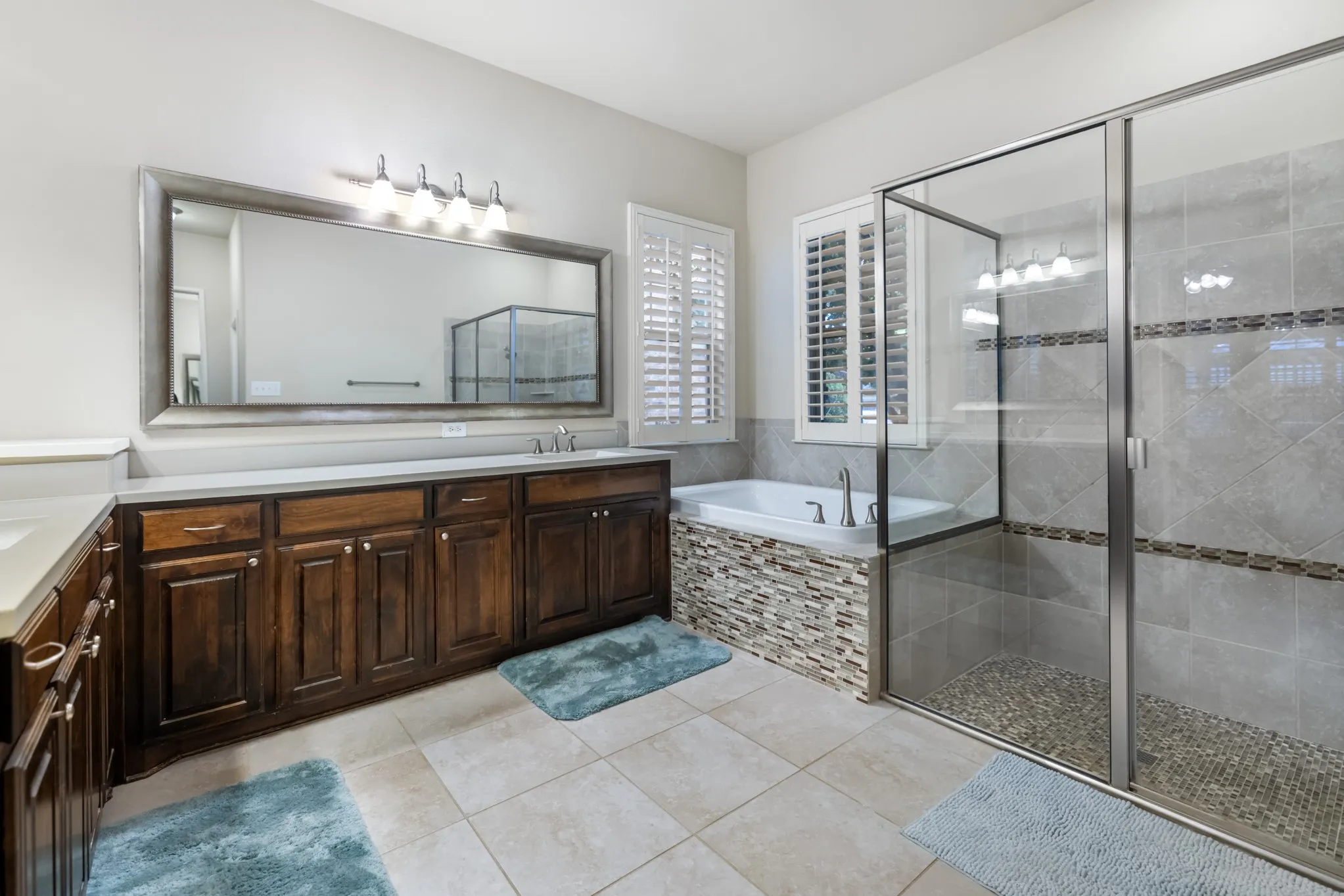 Spacious bathroom featuring a dual vanity with wood cabinetry and light countertops, a large mirror, a separate soaking tub with a stone accent surround, and a glass-enclosed shower with pebble flooring