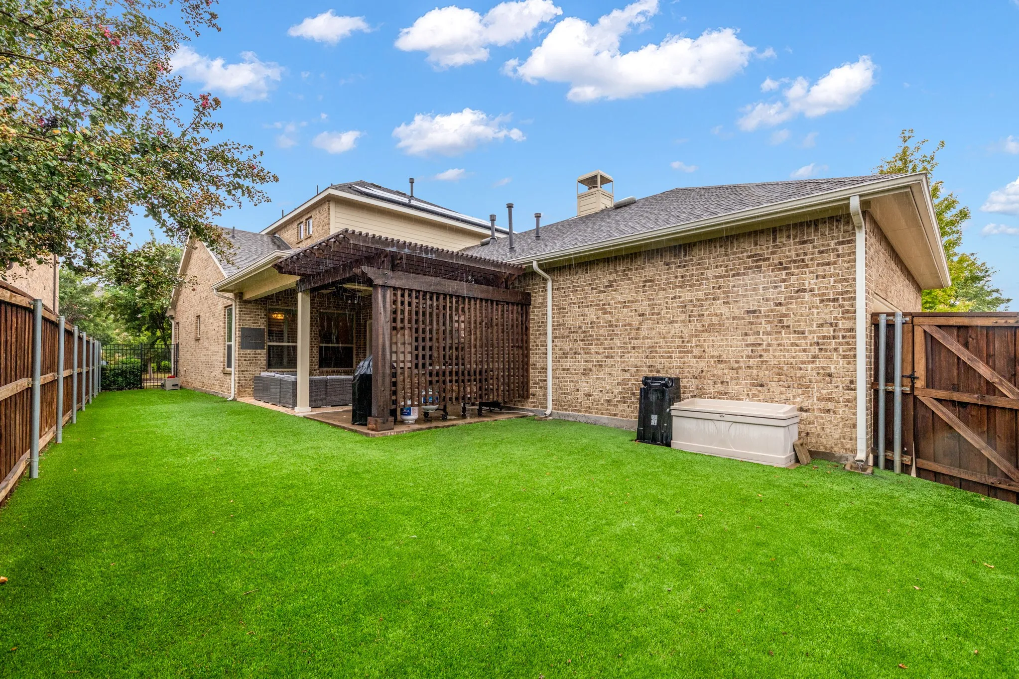 Expansive artificial turf backyard featuring a covered patio with a wooden pergola, brick exterior, and wood privacy fencing