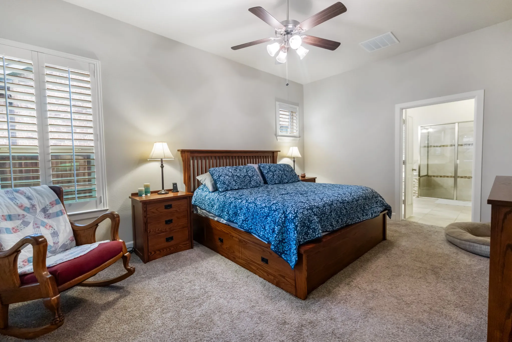 Carpeted bedroom featuring a ceiling fan, window with shutters, and an open doorway revealing a bathroom with a glass-enclosed shower