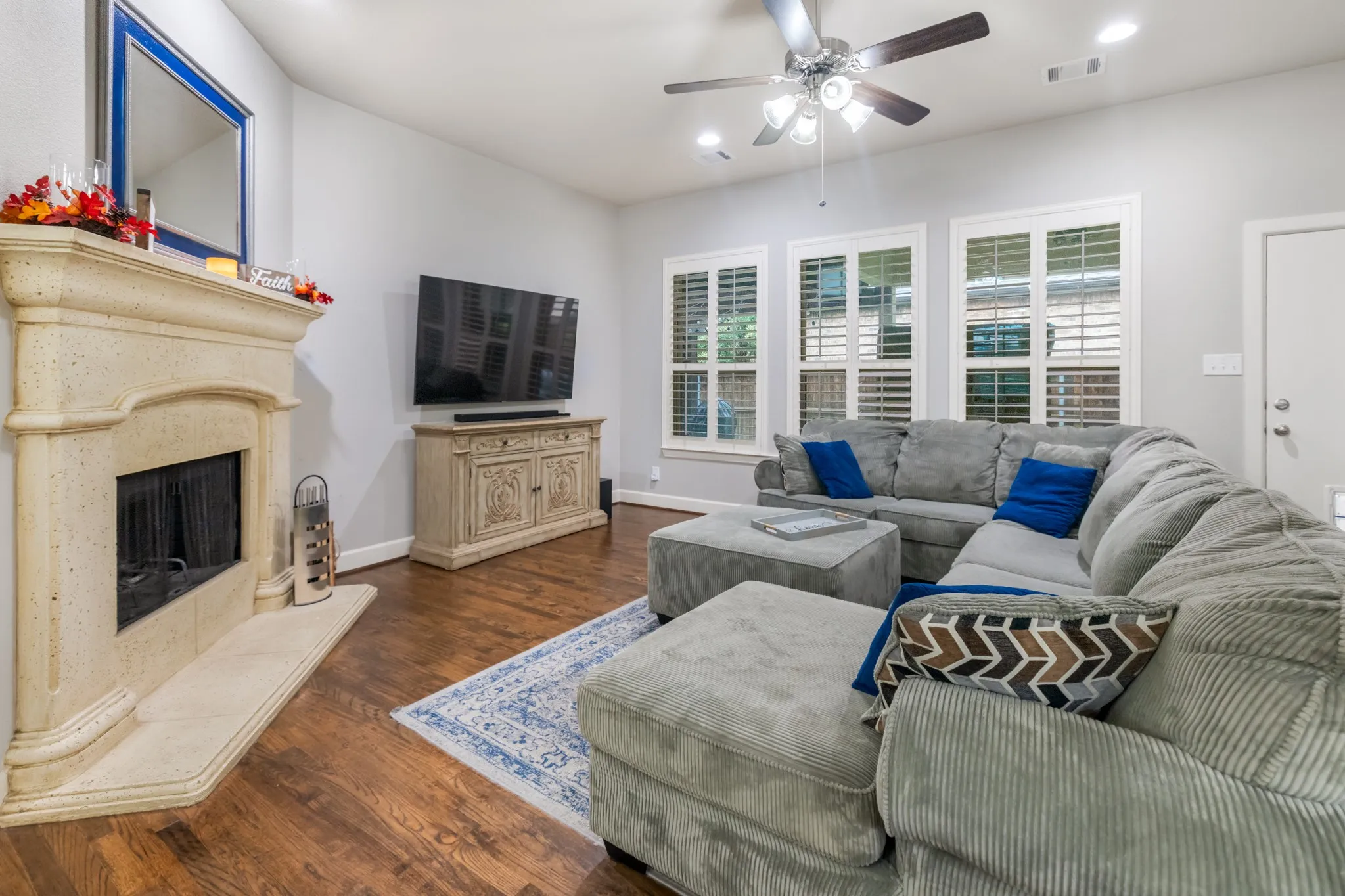 Wood-finish flooring, stone fireplace, multiple windows with plantation shutters, ceiling fan, and recessed lighting