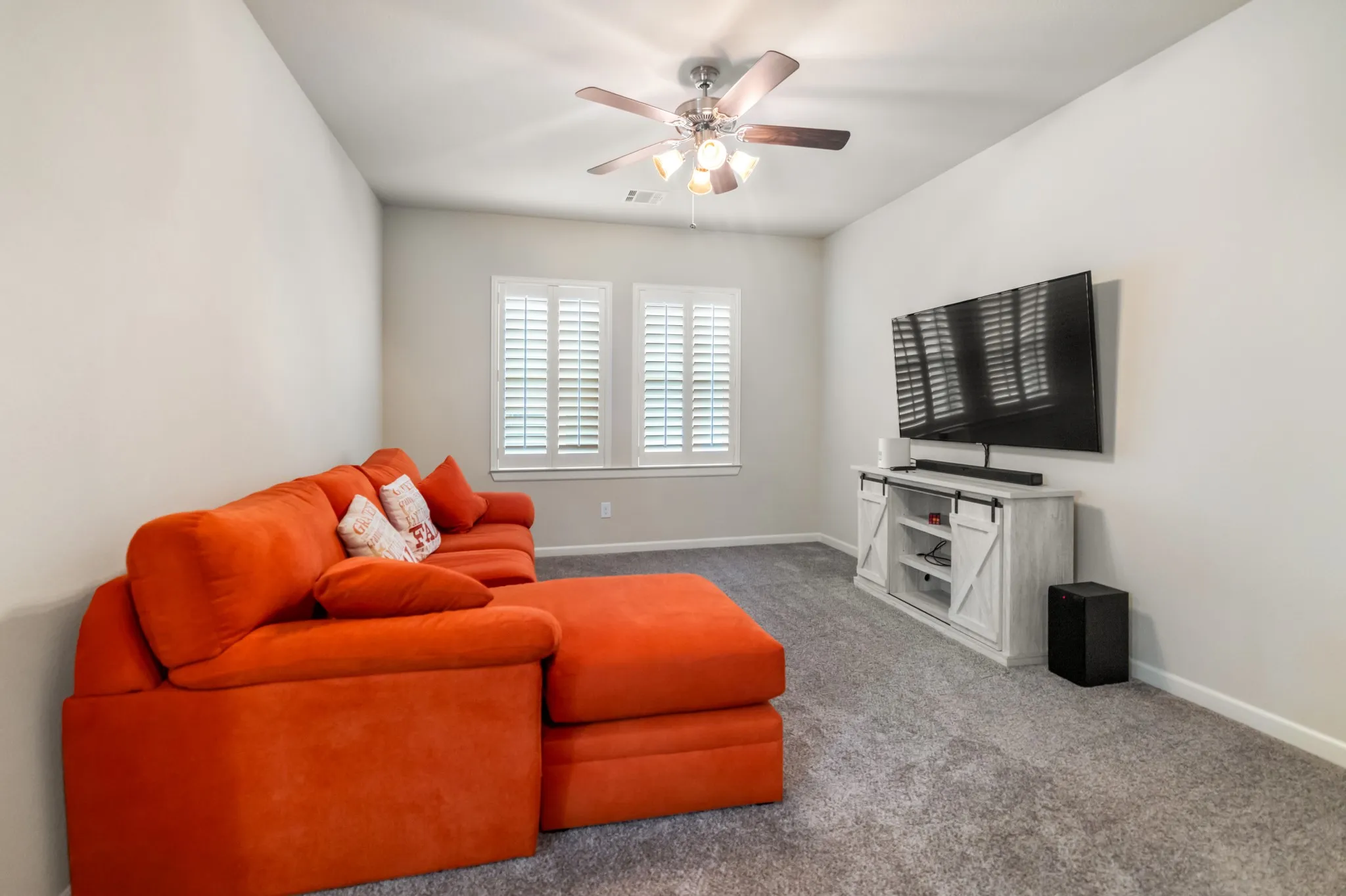 Upstairs.Wall-mounted television with a media console featuring sliding barn doors, two windows with plantation shutters, a ceiling fan with integrated lighting, and neutral carpet flooring
