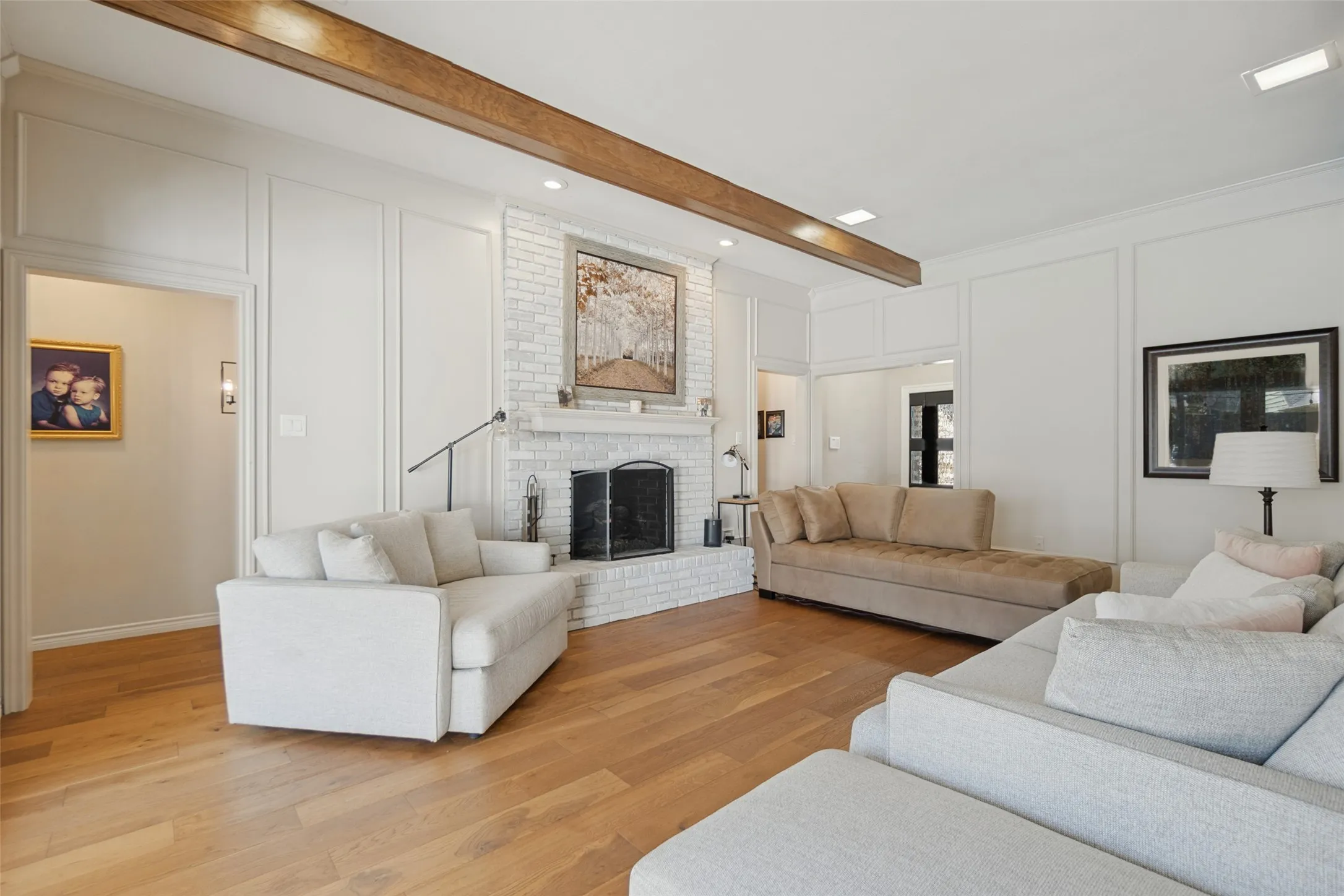 Spacious living area featuring wood-finish flooring, a white brick fireplace with a hearth, recessed lighting, decorative wall paneling, and an exposed ceiling beam