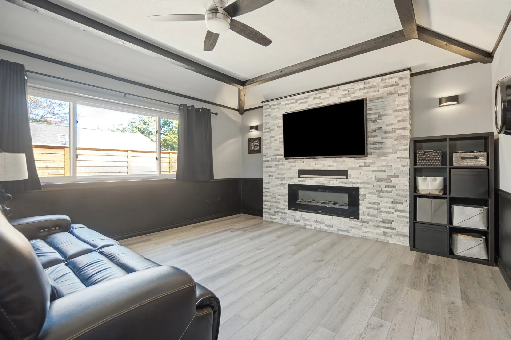 Light wood-finish flooring, accent wall with stacked stone-look tile, built-in electric fireplace, recessed wall lighting, and exposed ceiling beams