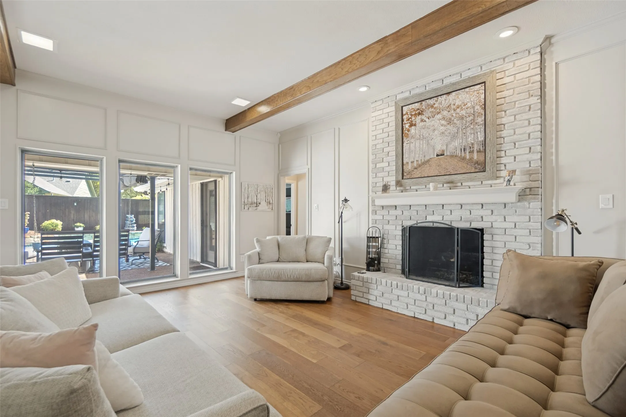 Hardwood flooring throughout, a white brick fireplace with a mantel, exposed wood beams, and a wall of glass doors opening to the exterior