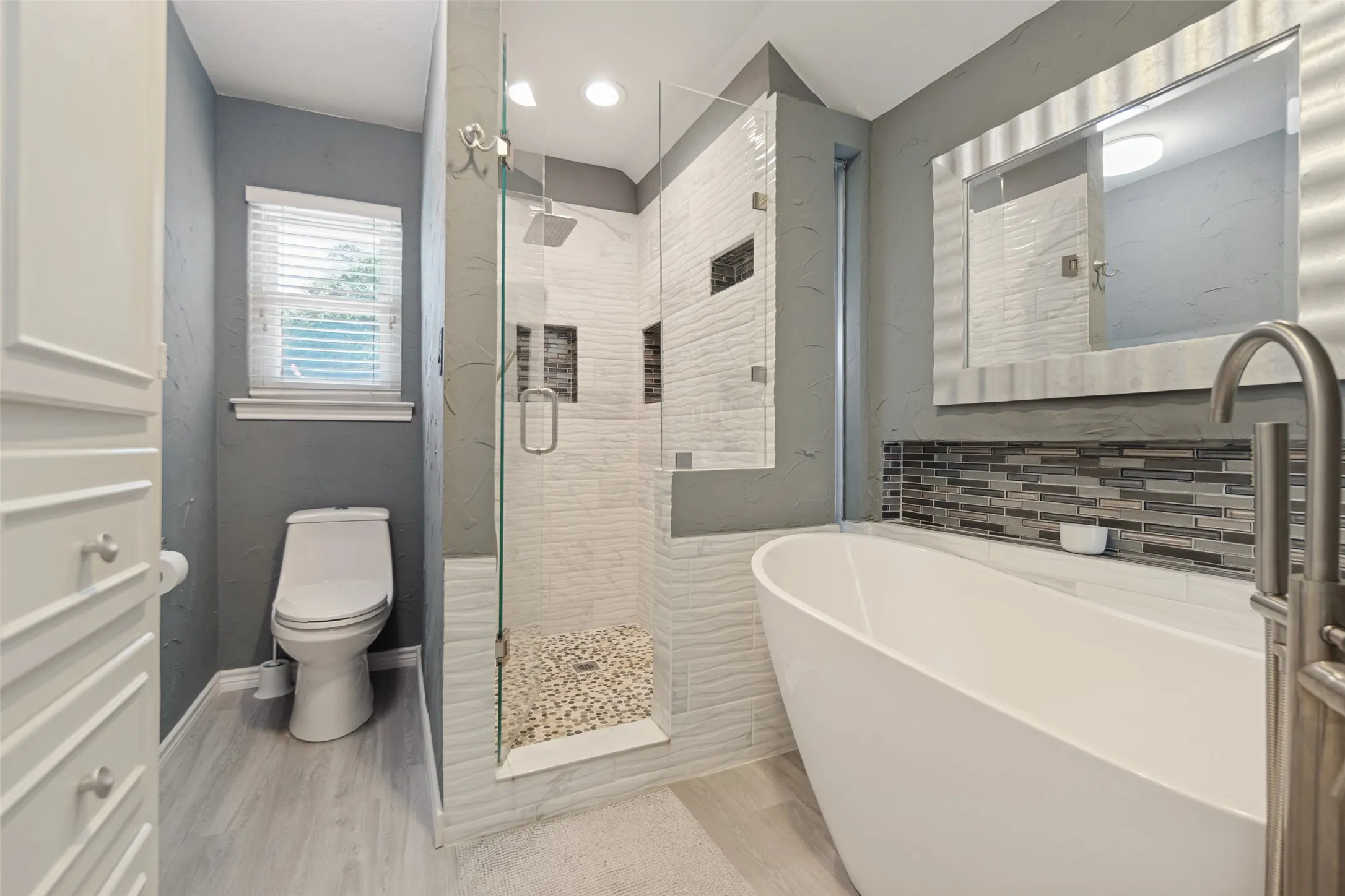 Freestanding soaking tub, glass-enclosed shower with pebble floor, rain showerhead, mosaic tile accent, and light wood-finish flooring
