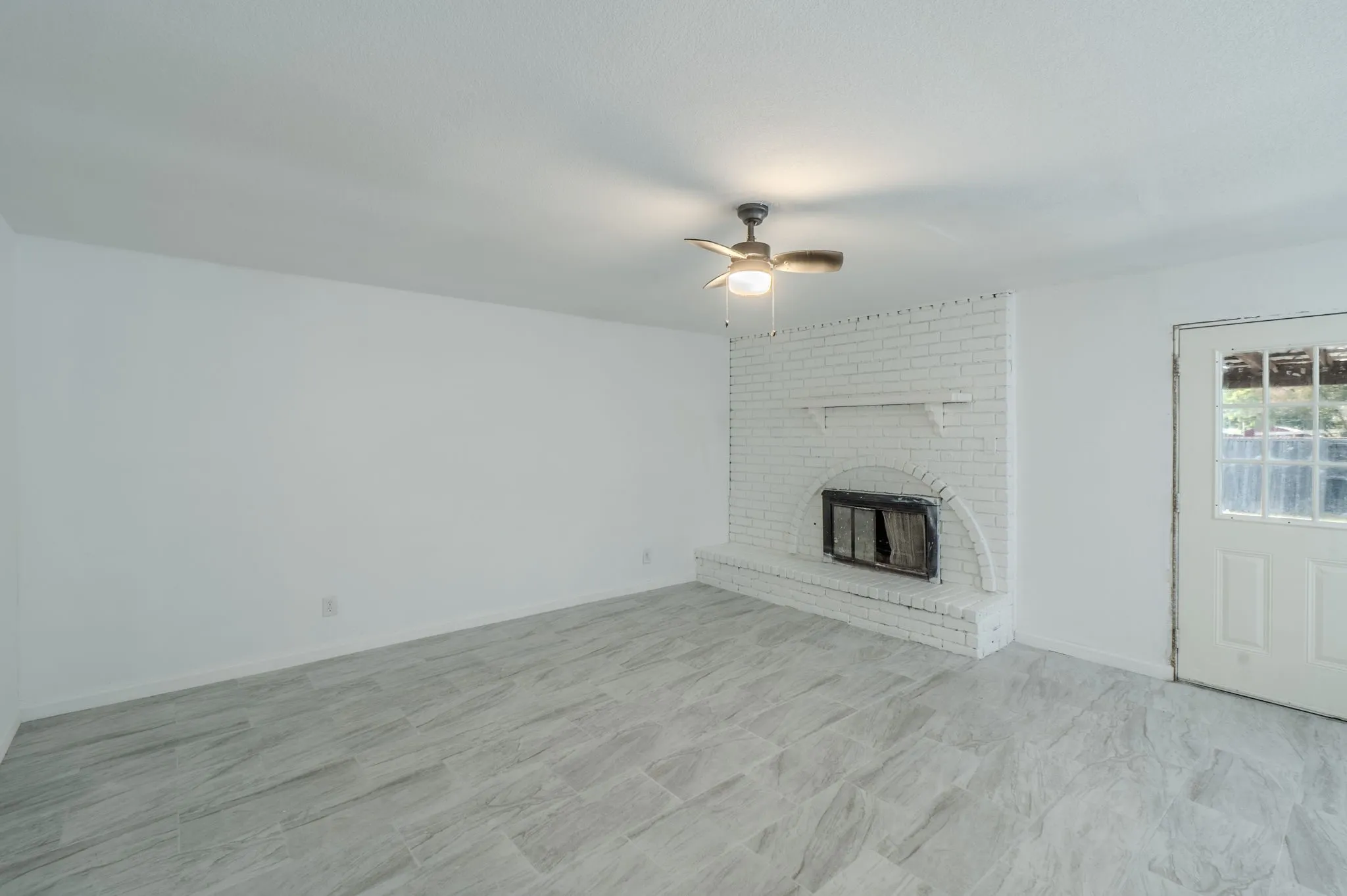 White brick fireplace with hearth, wood-finish ceiling fan with integrated lighting, and light grey tile flooring