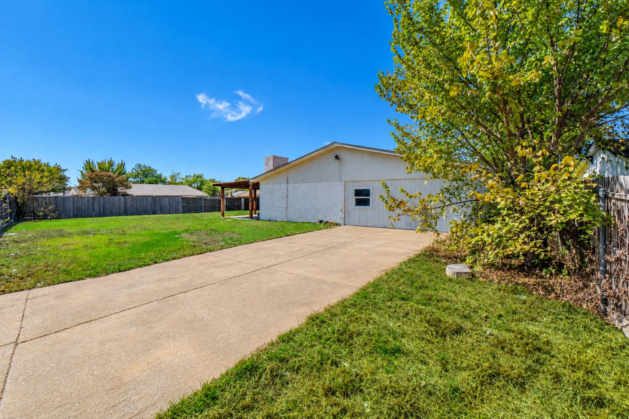 Expansive concrete driveway, attached garage, and a wood-fenced backyard featuring a pergola