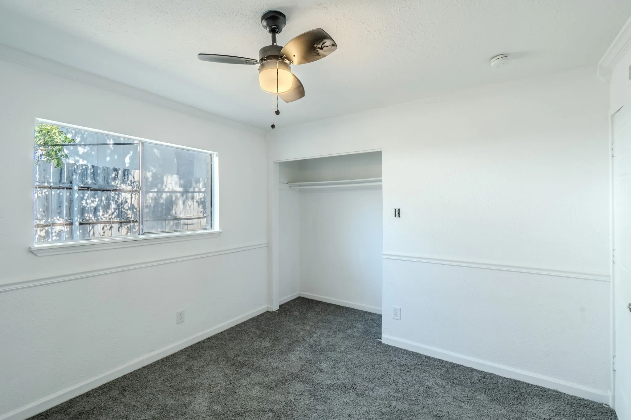 Recessed open closet with clothing rod, textured white walls, window with privacy screen, ceiling fan, and dark gray carpet