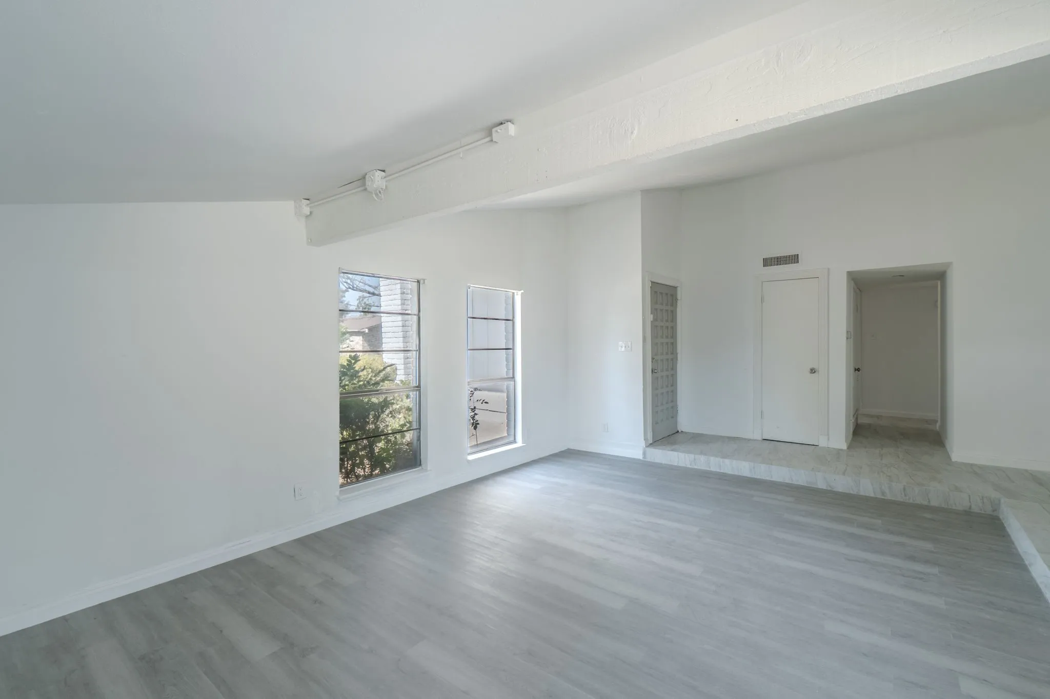 Spacious room with wood-finish flooring, white walls, exposed ceiling beam, multiple windows, and elevated tiled area