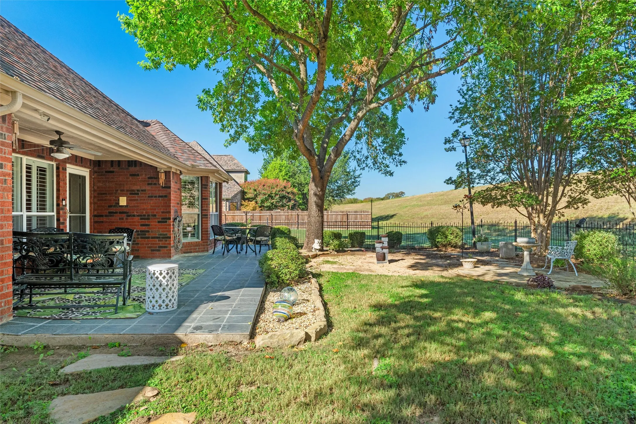 Brick exterior with multiple windows, covered patio with ceiling fan, stone-tiled patio, and a wrought iron fenced backyard with mature trees