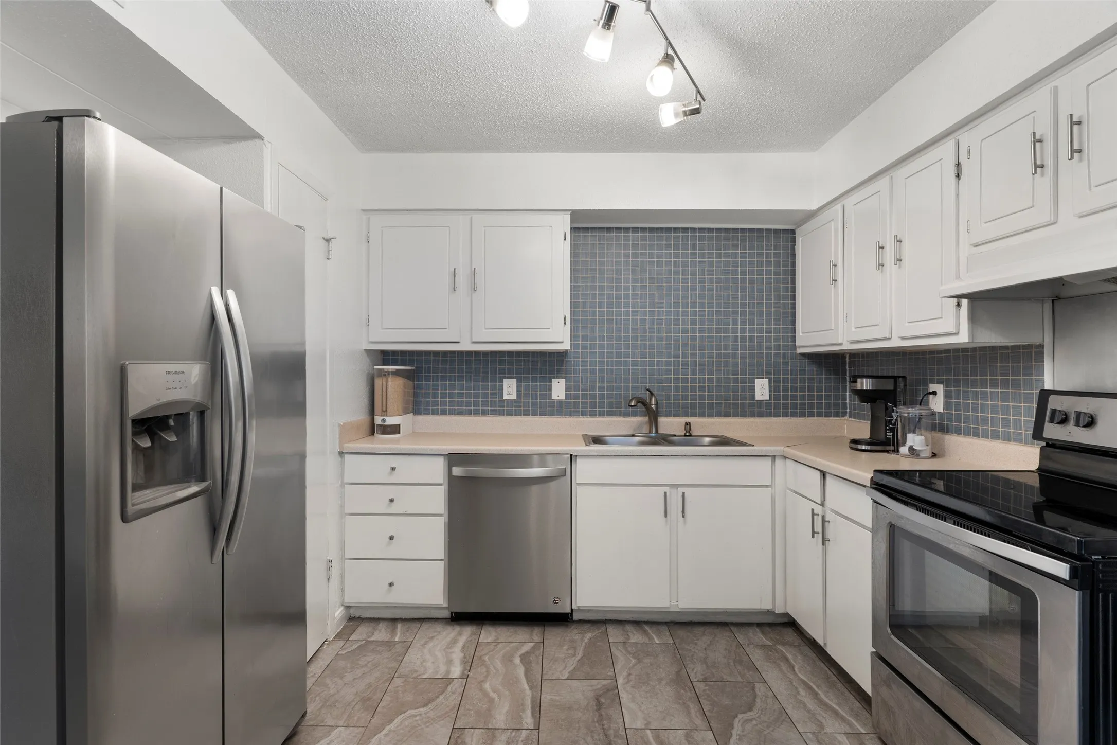 Modern kitchen featuring white cabinetry with chrome hardware, stainless steel appliances including a side-by-side refrigerator and dishwasher, blue subway tile backsplash, wood-finish flooring, and an L-shaped layout with ample counter and storage space