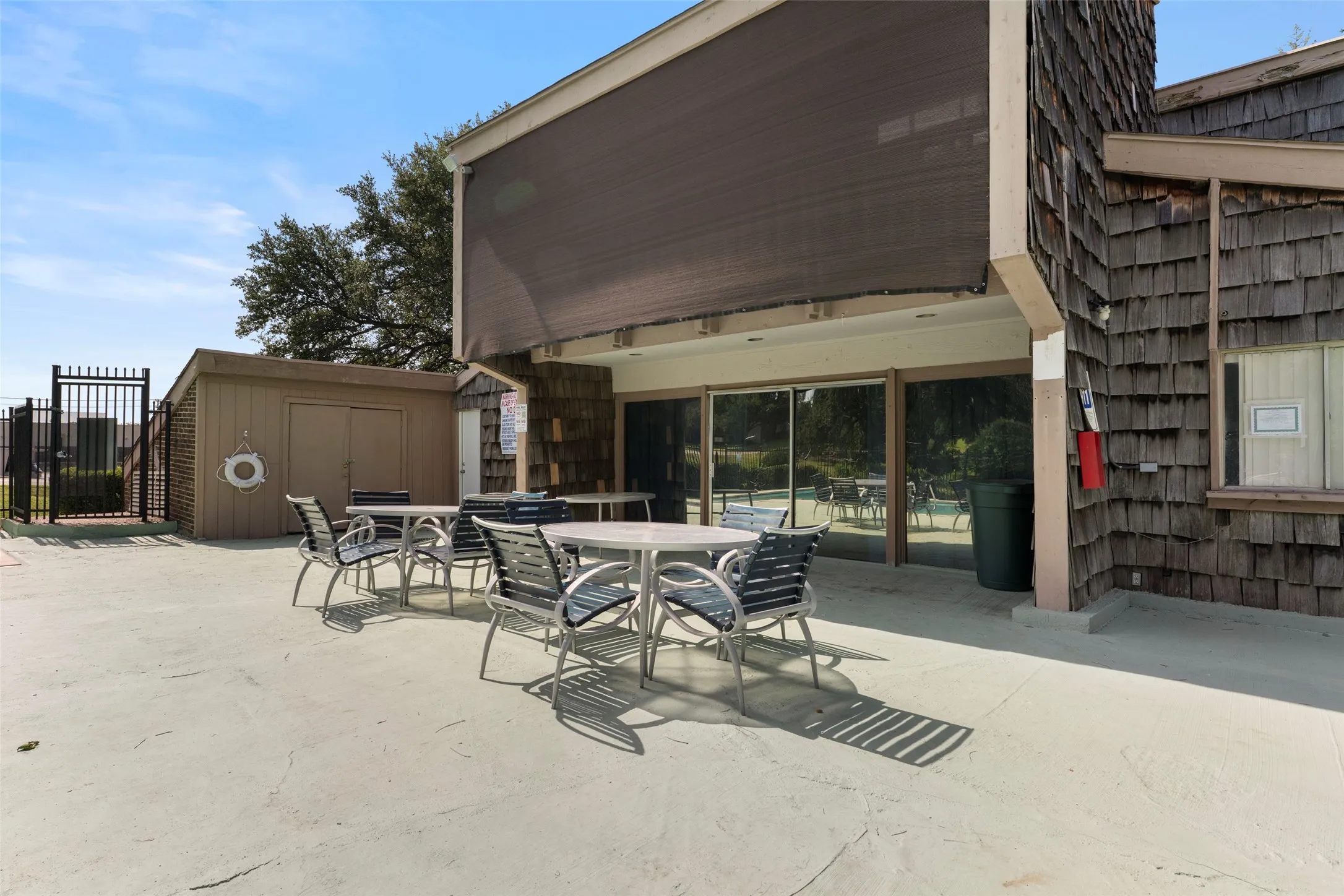 Outdoor patio area with an overhead sunshade, wood shingle siding, an adjacent storage shed, and a black wrought iron fence