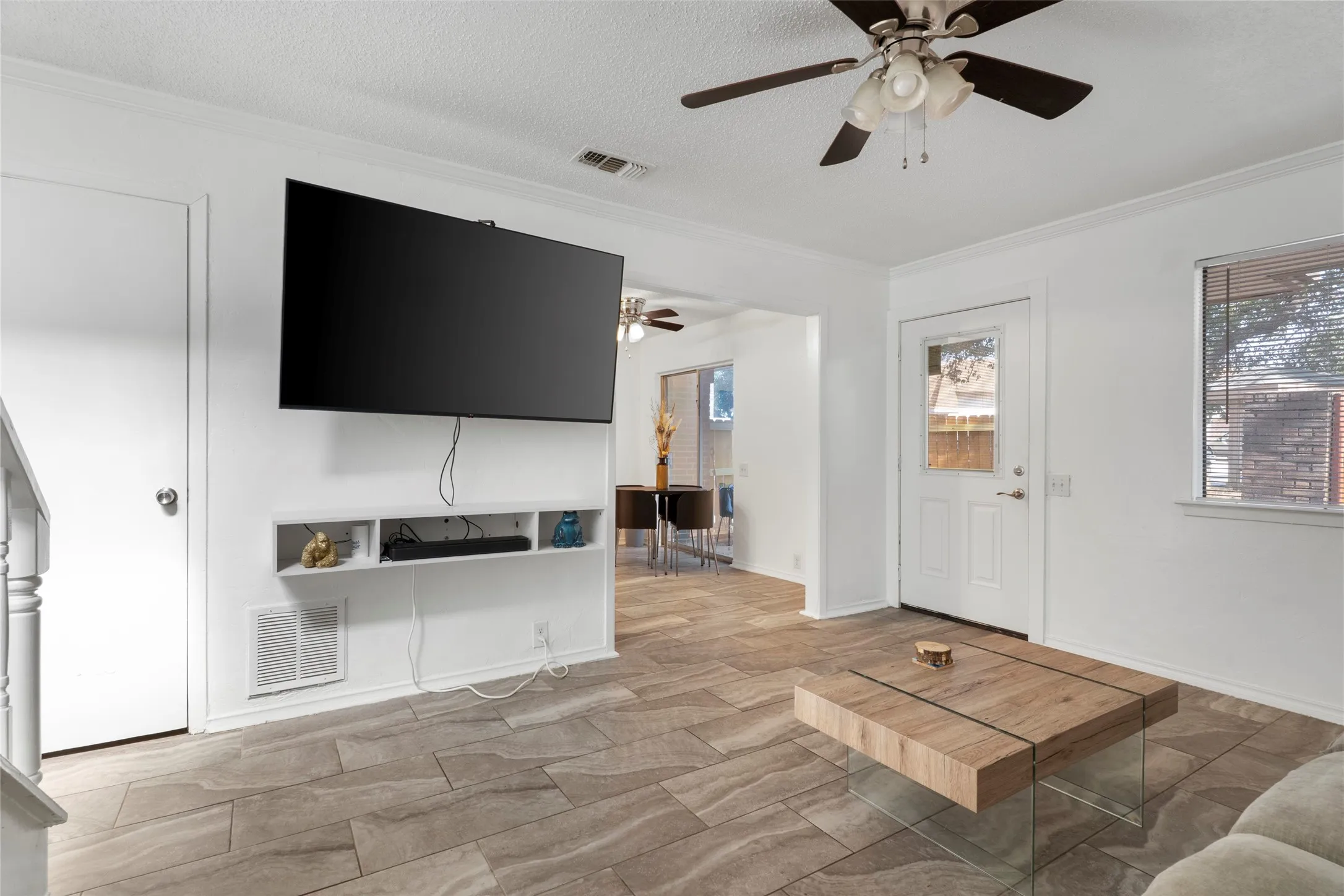 Spacious living area featuring a wall-mounted flat-screen television with built-in shelving, wood-finish plank flooring throughout, ceiling fan with integrated lighting, and multiple entry points