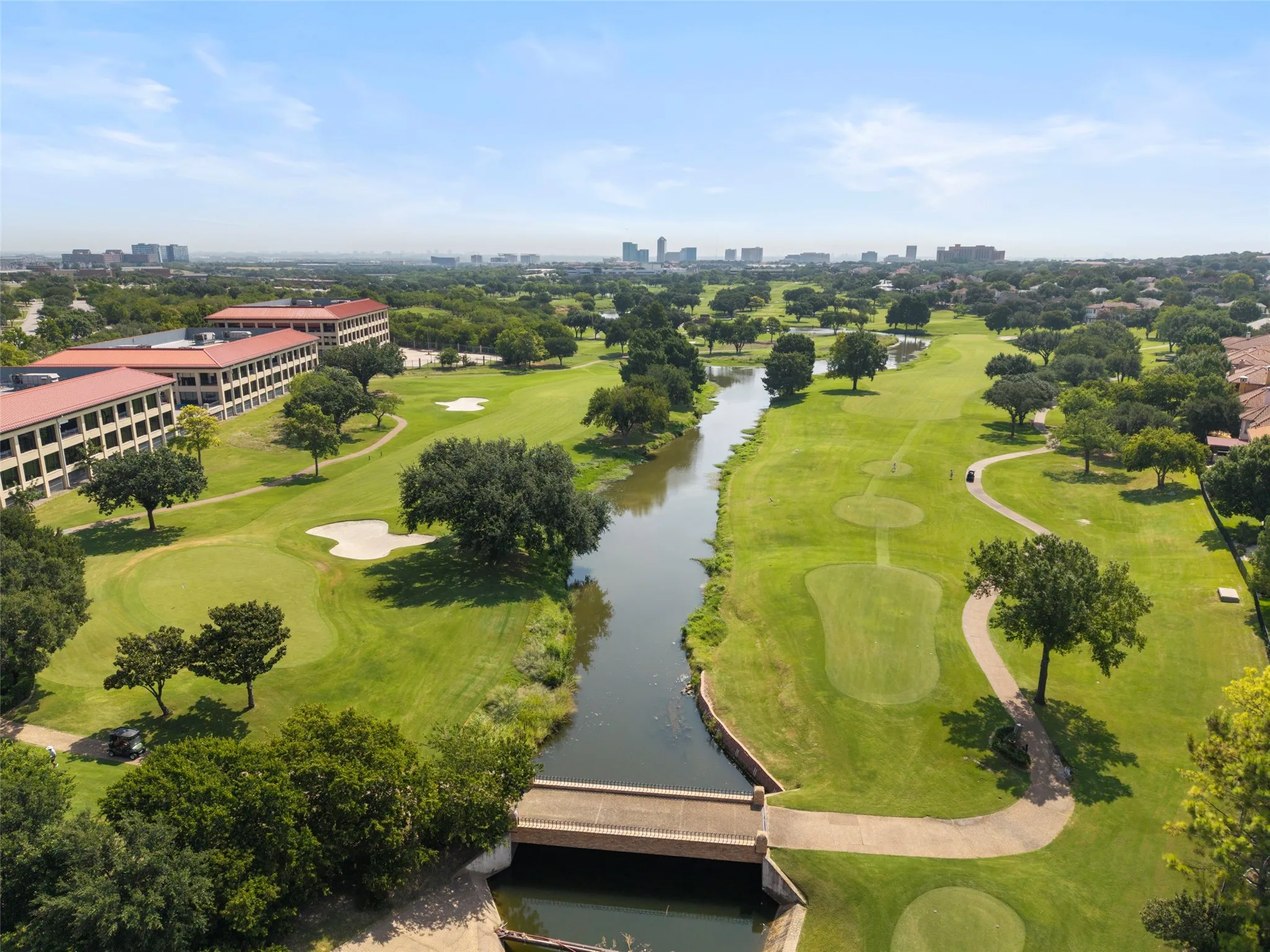 Expansive green golf course with a water feature, mature trees, and a distant city skyline