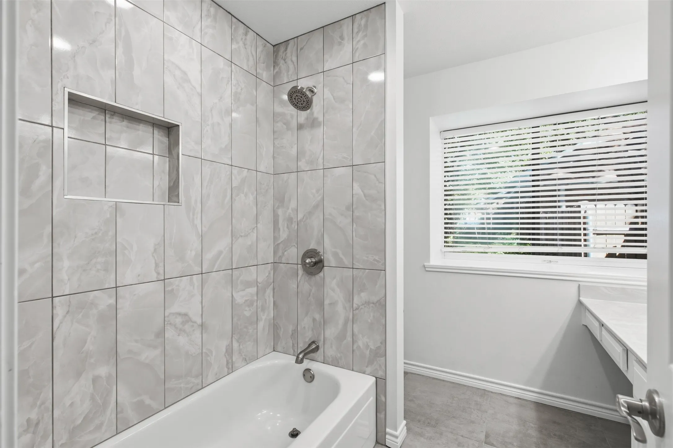 Bathtub with grey tiled surround, built-in shower niche, and brushed nickel fixtures