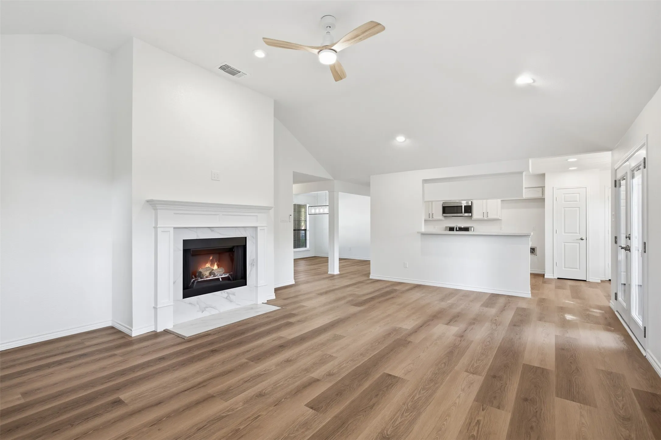 Vaulted ceiling with a ceiling fan, fireplace with a marble-look surround, wood-finish flooring, kitchen pass-through with white cabinetry, and double glass doors