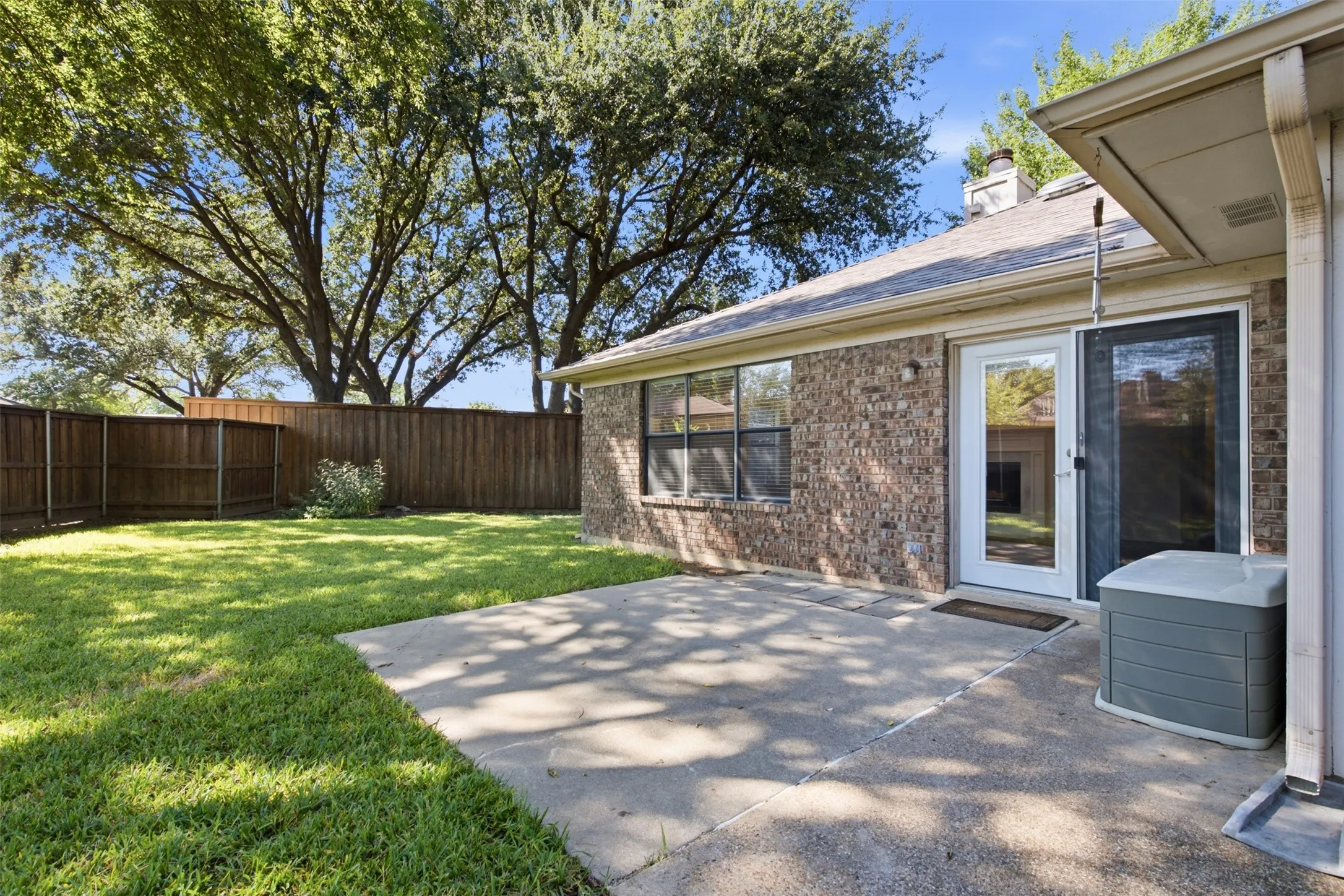 Brick exterior with expansive concrete patio overlooking a manicured lawn anchored by mature shade trees