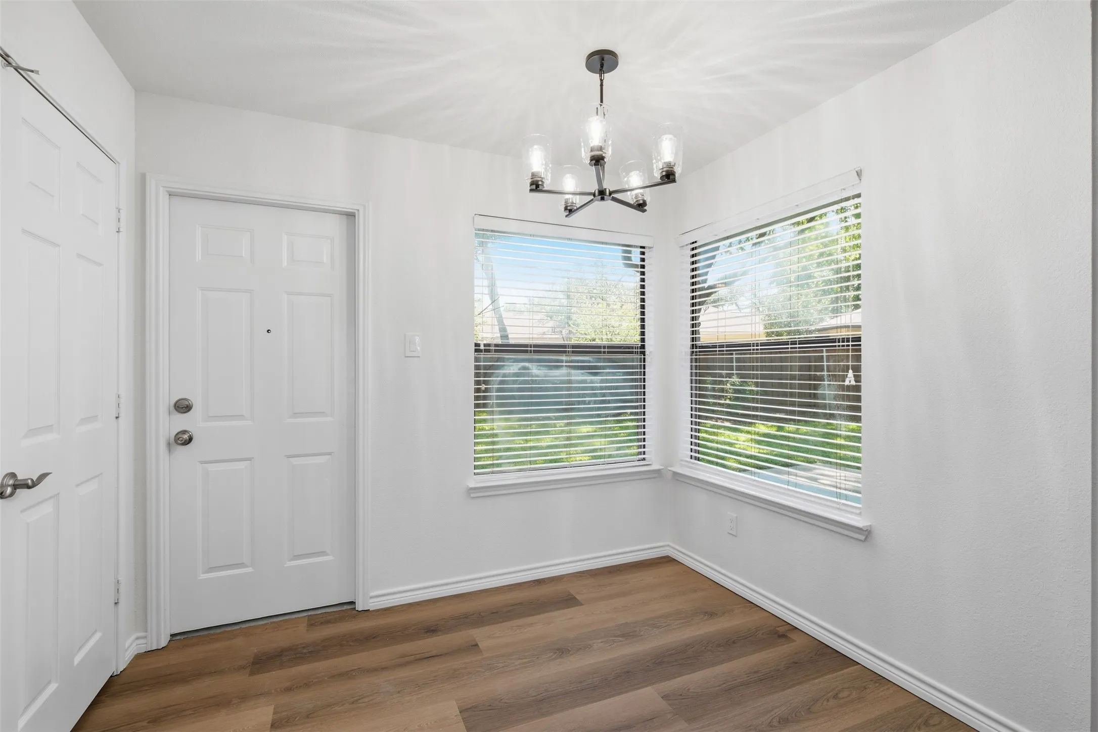 White paneled door, wood-finish flooring, two windows with blinds, and a modern chandelier