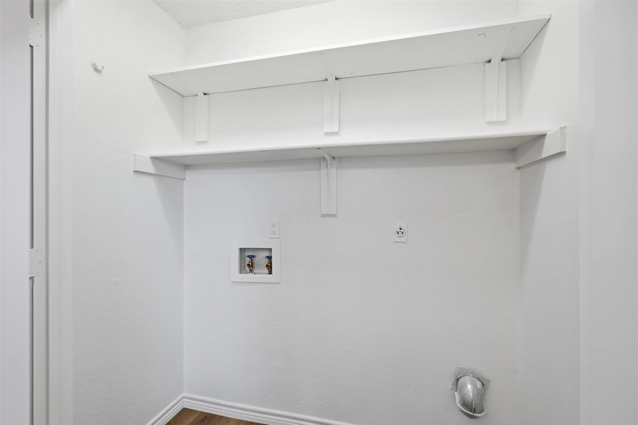 White interior walls with two white wall-mounted shelves, wood-finish flooring, and utility hookups