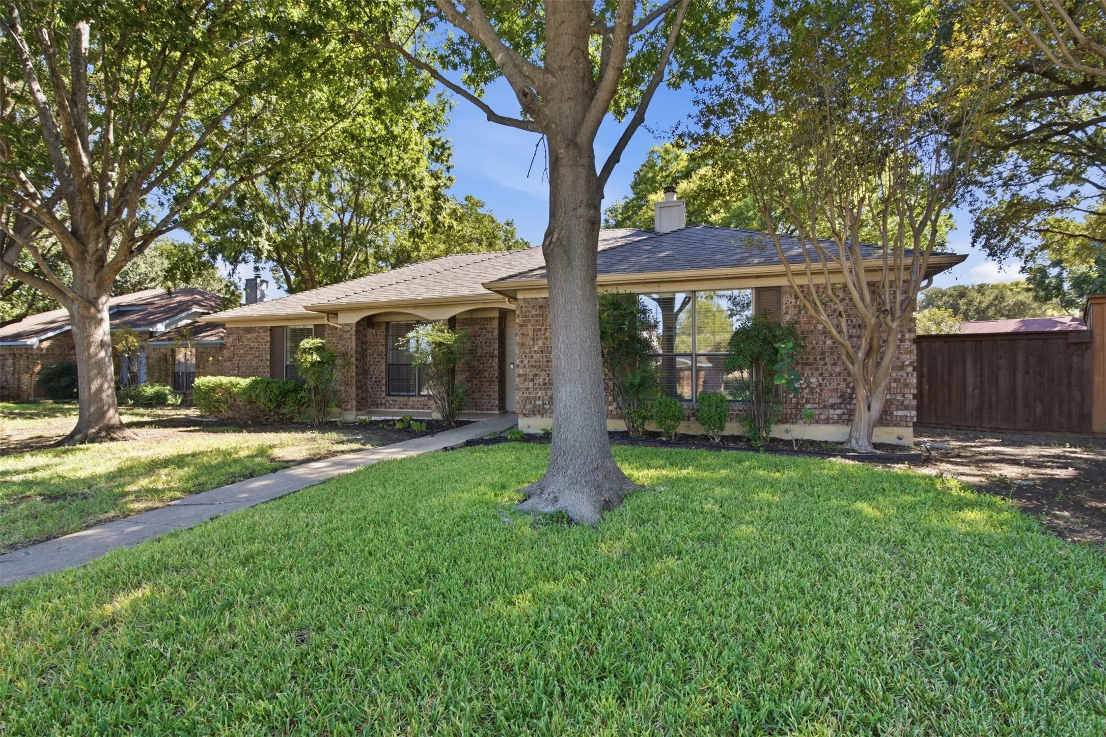 Mature shade trees frame this expansive lawn with concrete walkways, brick exterior construction, and covered porch areas with beam detailing